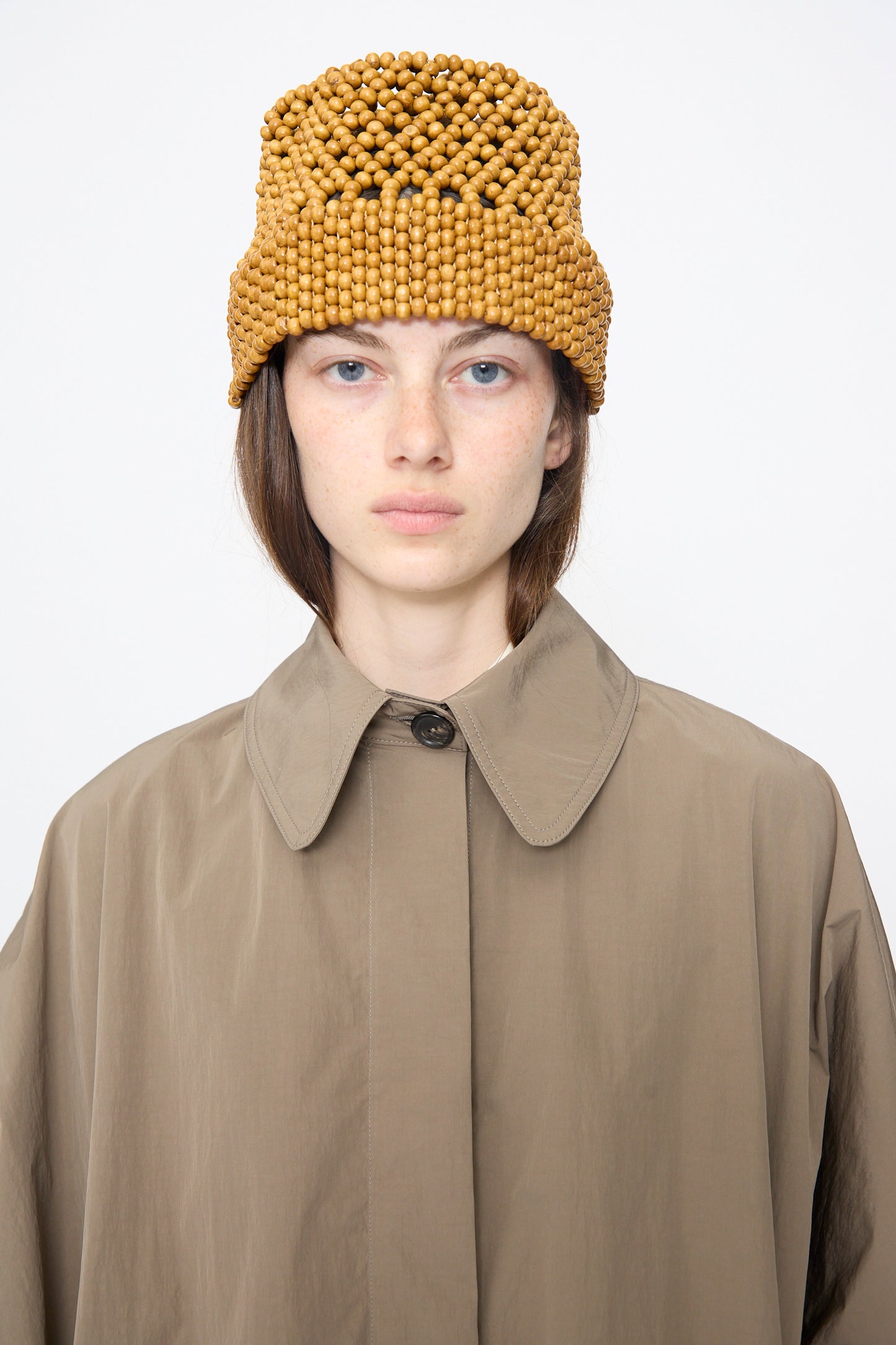 A person wearing the Cordera Batwing Trench in Vetiver, a tan cotton-blend oversized coat, and a brown beaded hat, facing forward against a plain white background.