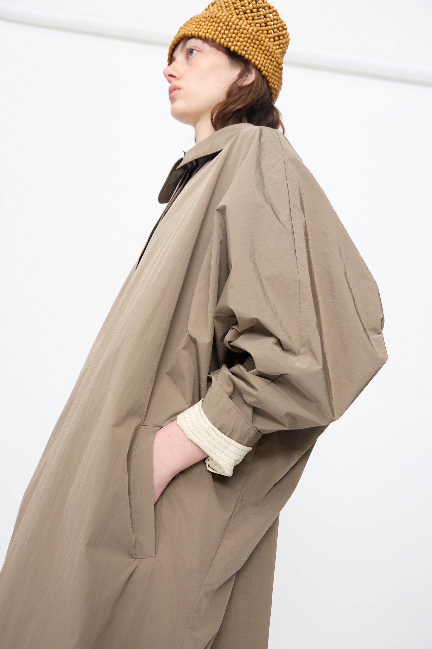 A person wears Cordera's Batwing Trench in Vetiver—a water-repellent, oversized taupe coat—paired with a long-sleeve shirt with rolled cuffs and a textured brown knit hat, standing against a plain white background.