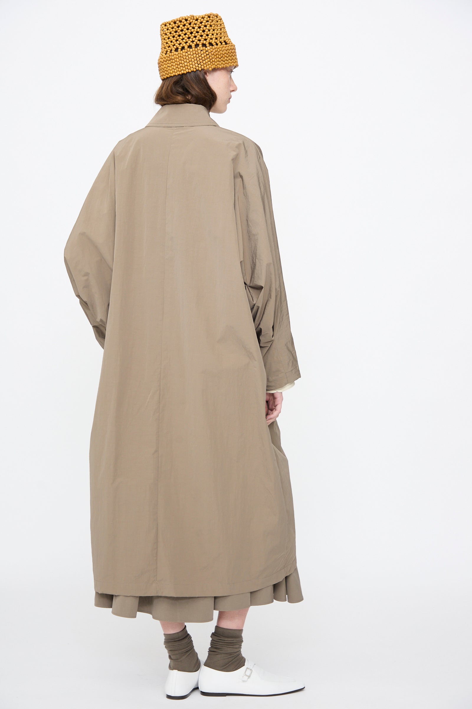 A person stands with their back to the camera against a plain white background, wearing Cordera's Batwing Trench in Vetiver, a cotton blend oversized coat, paired with a yellow knit hat, grey socks, white shoes, and a pleated skirt.