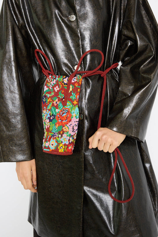 A person wearing a black leather coat holds a red drawstring pouch, with a handmade Beaded Bag in Flores by Cordera adorned with a floral pattern secured around their waist.