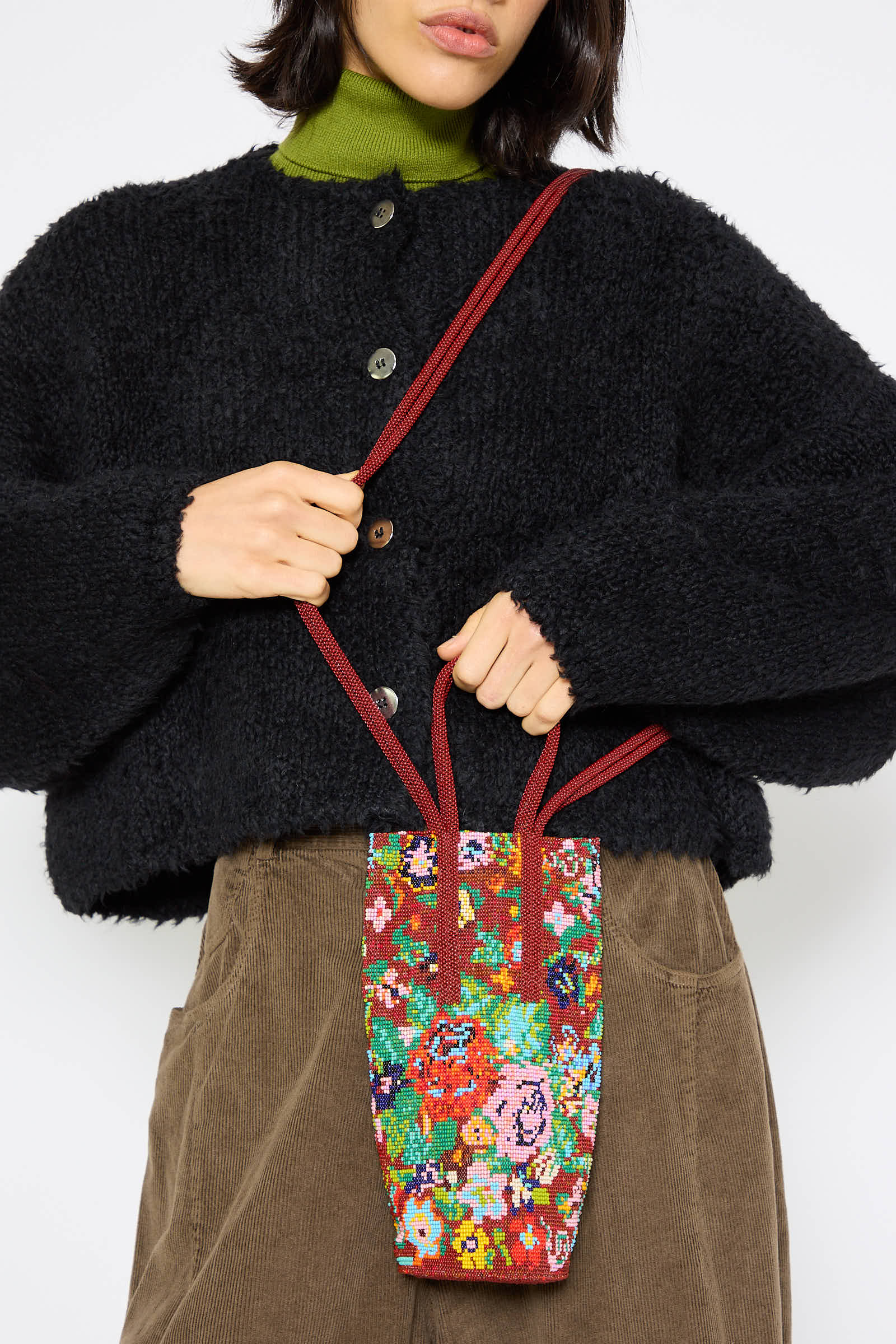 A person wearing a black fuzzy cardigan and a green top is holding the Cordera Beaded Bag in Flores, which features red straps and is adorned with a flower motif.