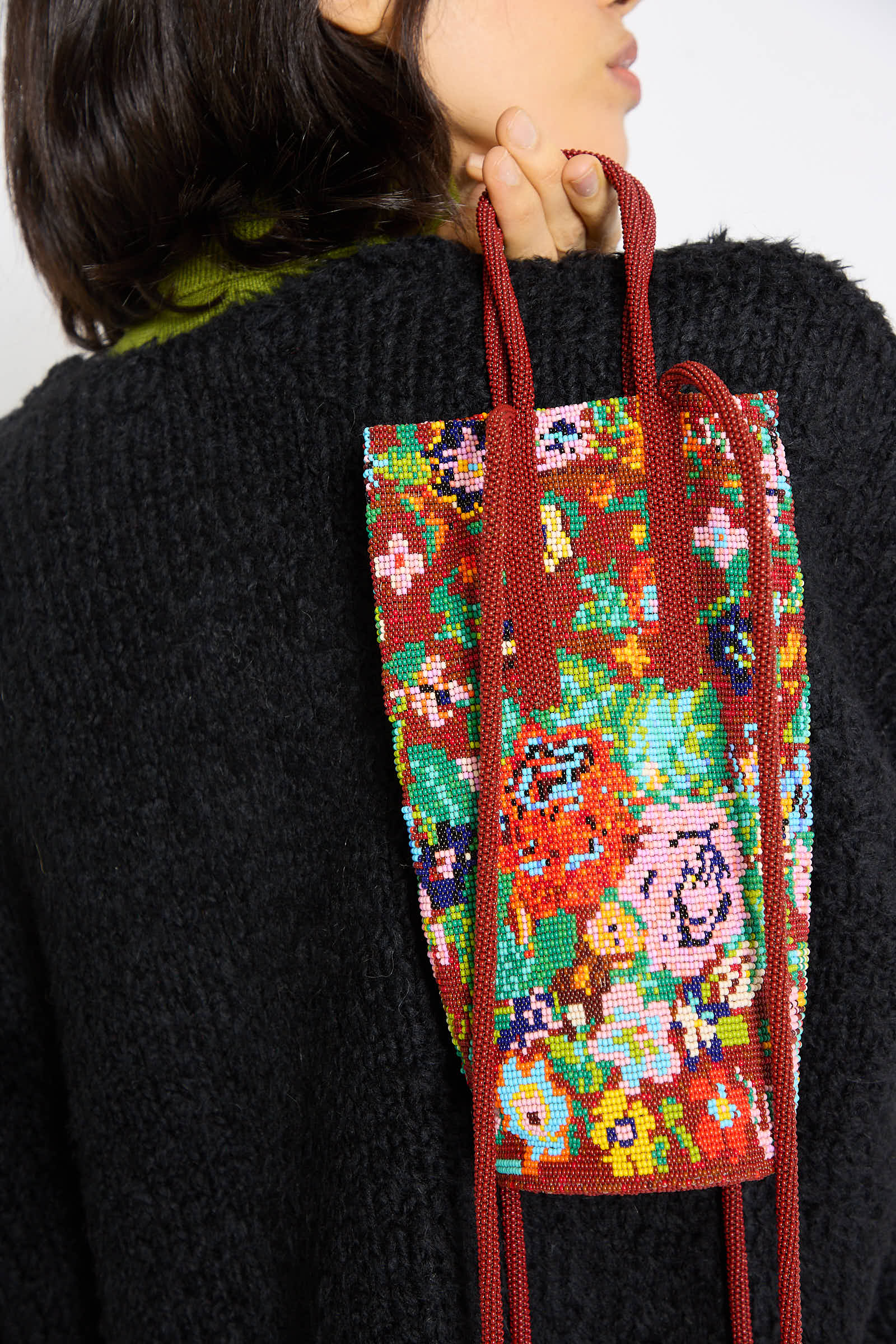 A person with dark hair is wearing a black sweater and holding the Cordera Beaded Bag in Flores, which features multicolored embroidery and beaded drop handles, draped over their shoulder.