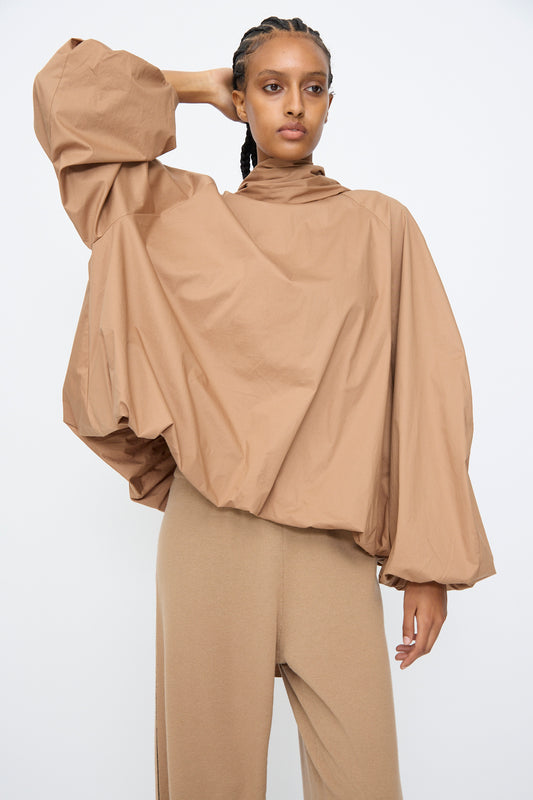 A person stands against a plain background wearing Cordera’s Bow Shirt in Camel, a loose cotton-blend top with voluminous sleeves, and matching tan pants, one arm bent with a hand touching their head.