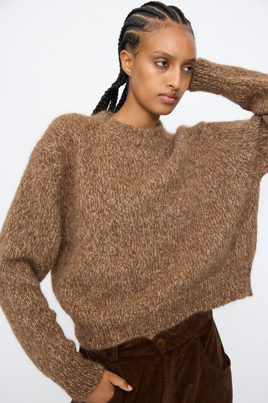 Wearing Cordera's Chunky Sweater in Brown Melange and brown corduroy pants, a person with braided hair poses with one arm raised behind their head against a plain background.