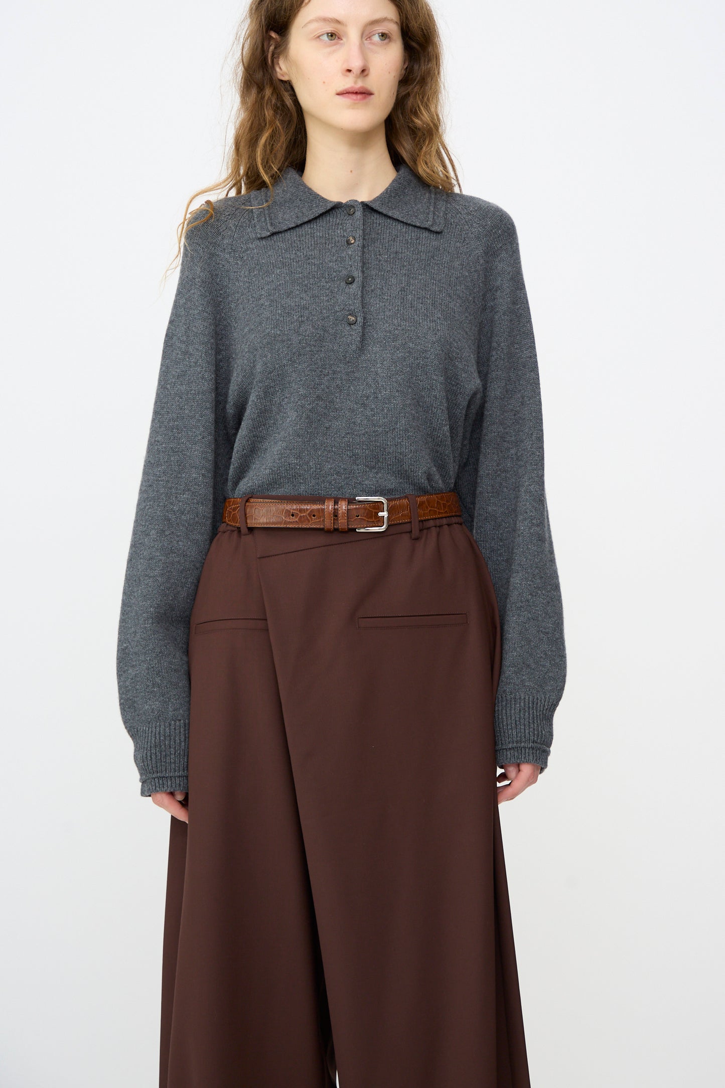 A person wears a grey long-sleeve collared sweater tucked into high-waisted, wide-leg brown pants, accessorized with the Cordera Classic Belt in Caramel Crocodile, standing against a plain white background.