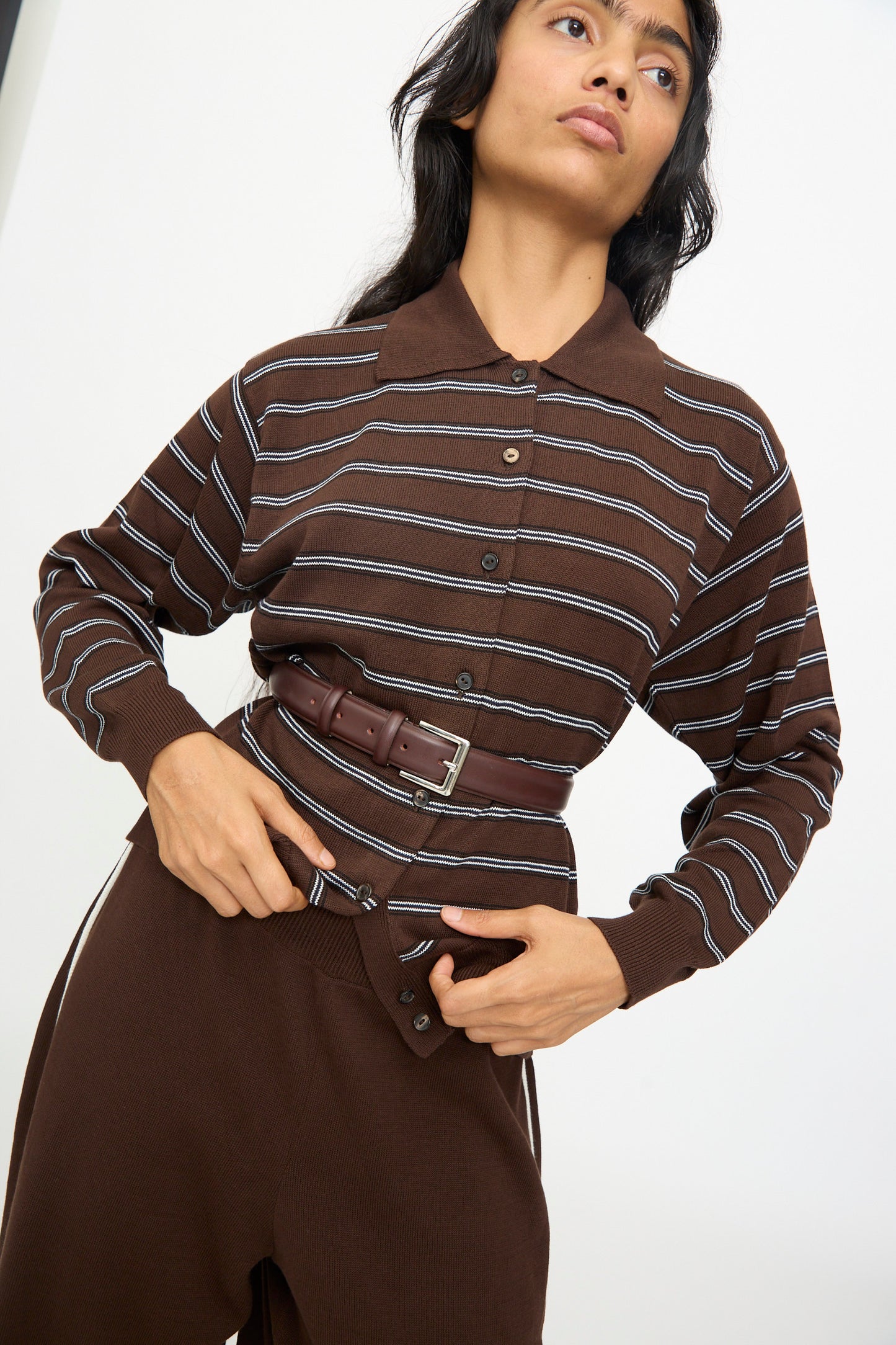 A person in a brown, long-sleeve, collared shirt with white stripes is wearing Cordera's Classic Belt in Prune, crafted from calfskin leather in Spain, over the shirt and matching brown pants, which are partially unbuttoned at the waist.