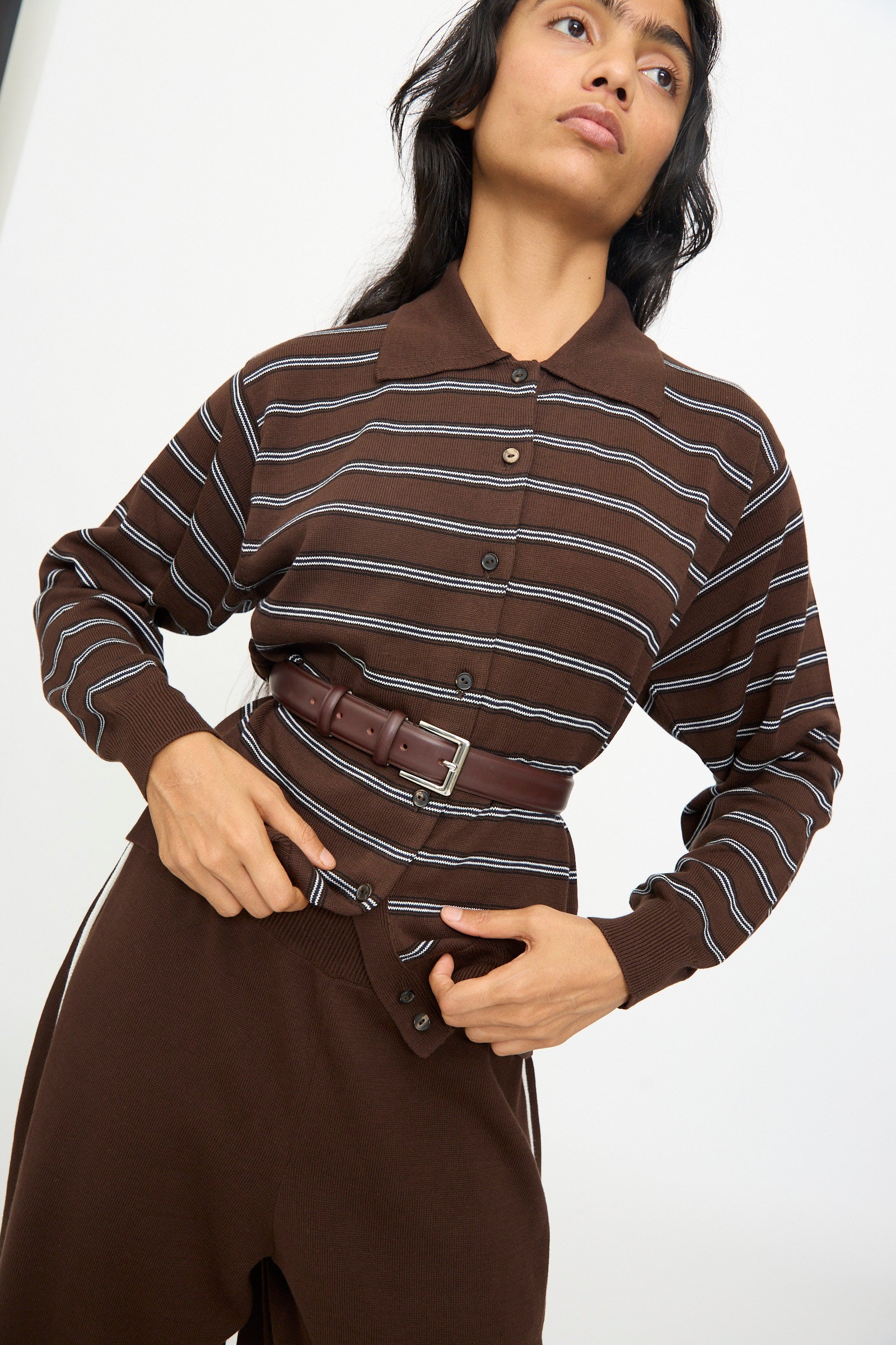 A person in a brown, long-sleeve, collared shirt with white stripes is wearing Cordera's Classic Belt in Prune, crafted from calfskin leather in Spain, over the shirt and matching brown pants, which are partially unbuttoned at the waist.