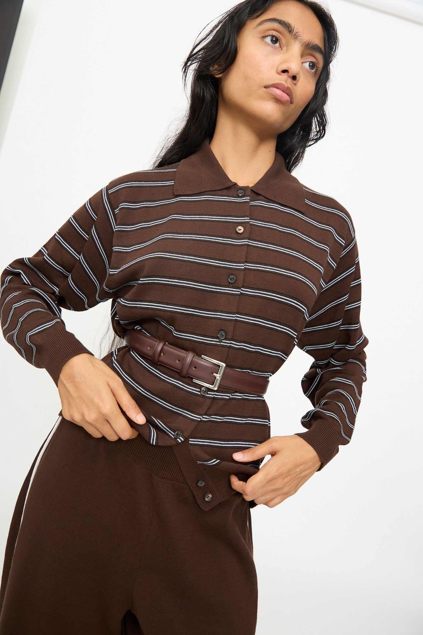 A woman wearing a brown button-up shirt with white stripes, brown high-waist pants, and the Cordera Classic Belt in Prune—crafted from calfskin leather and finished with a silver buckle—stands against a plain background.