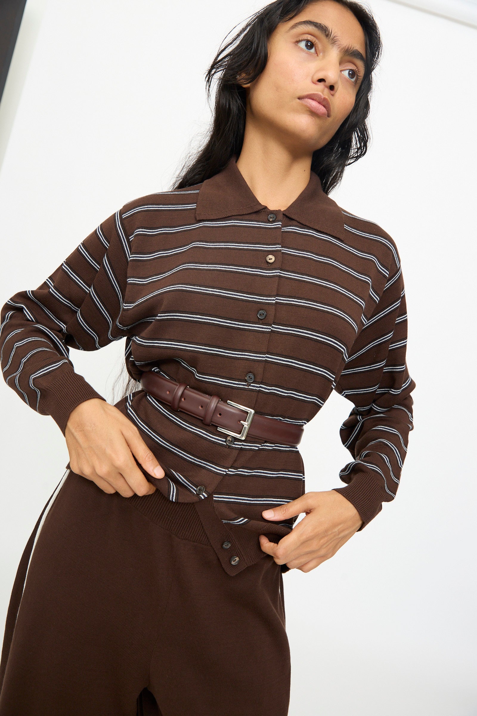 A woman wearing a brown button-up shirt with white stripes, brown high-waist pants, and the Cordera Classic Belt in Prune—crafted from calfskin leather and finished with a silver buckle—stands against a plain background.