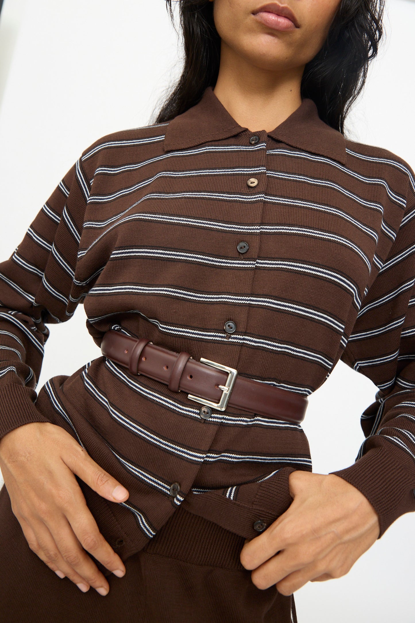 A person wears a brown striped button-up shirt with the Cordera Classic Belt in Prune, crafted from calfskin leather in Spain, fastened tightly around the waist over the shirt.