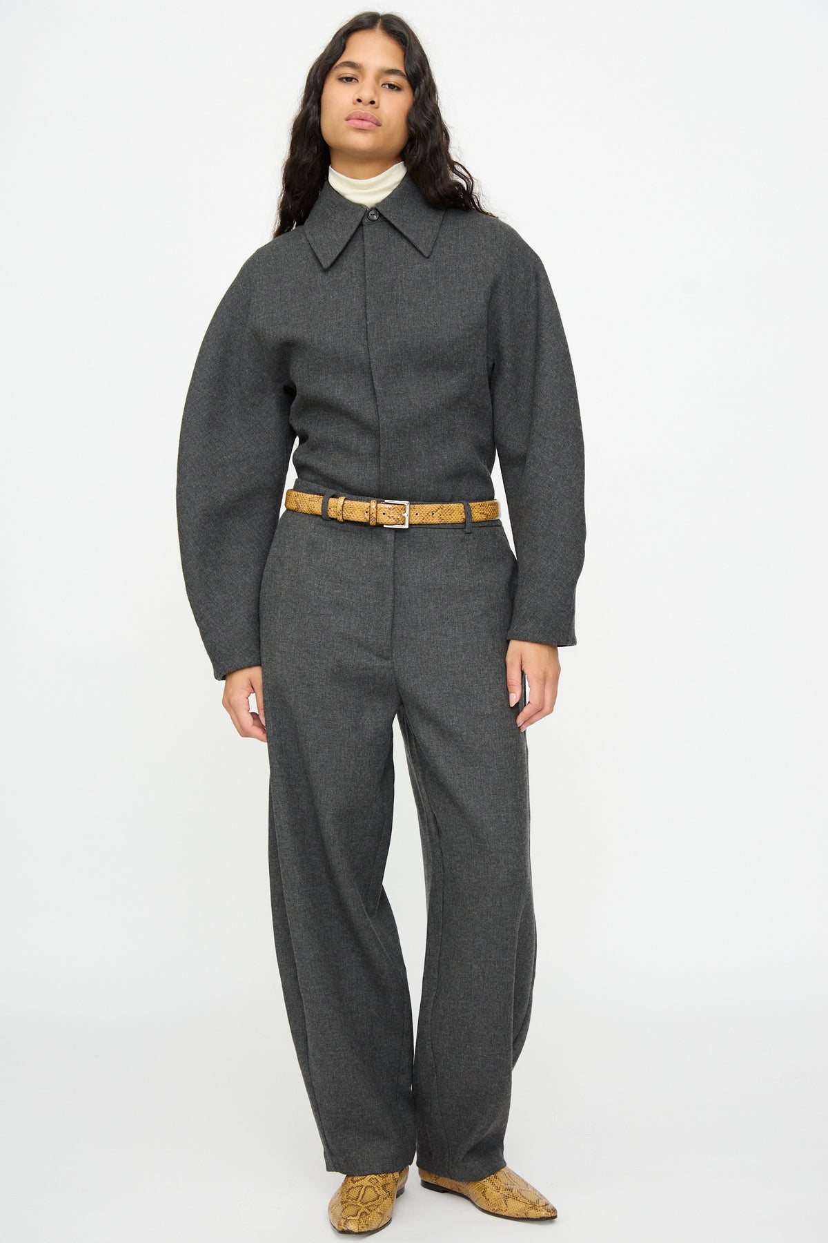 A person wearing a grey long-sleeved jumpsuit cinched at the waist with a tan Classic Belt in Snake, paired with matching snakeskin-print boots.