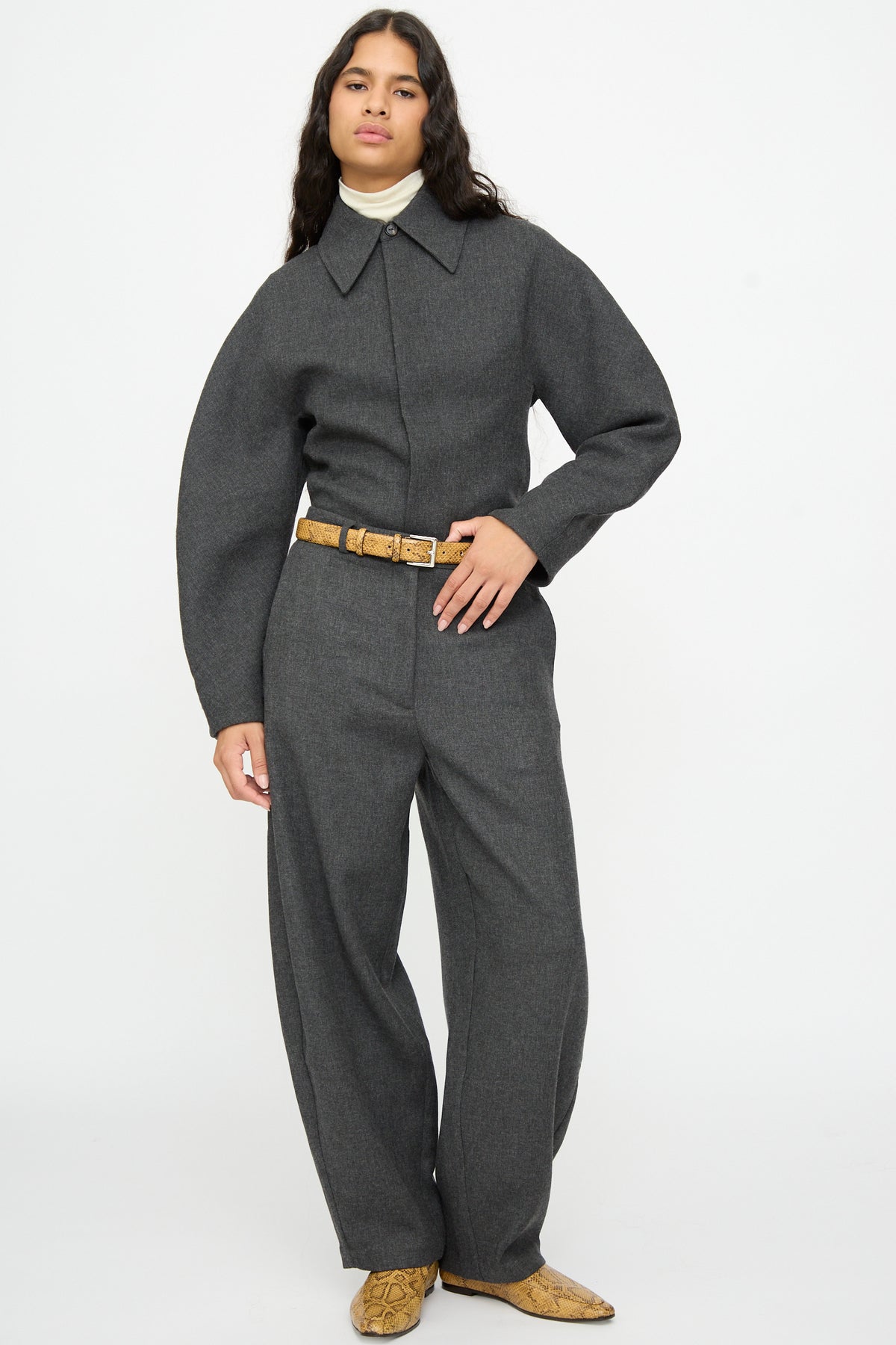 A model wears a gray collared jumpsuit cinched at the waist with a tan snake print Classic Belt in Snake by Cordera, paired with matching snakeskin-patterned boots.