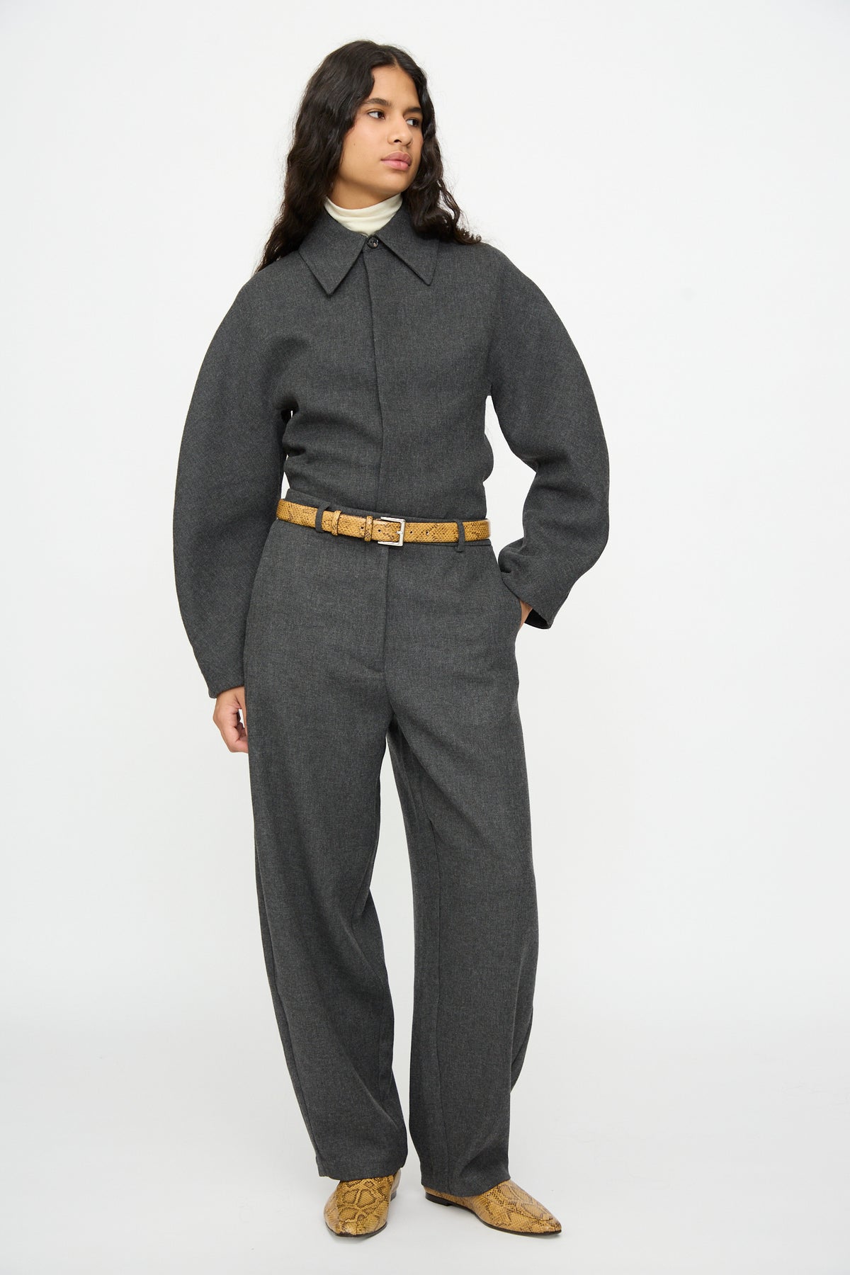 A person wears a charcoal grey jumpsuit, a tan snakeskin-print Classic Belt in Snake, and matching snakeskin-print pointed-toe shoes.