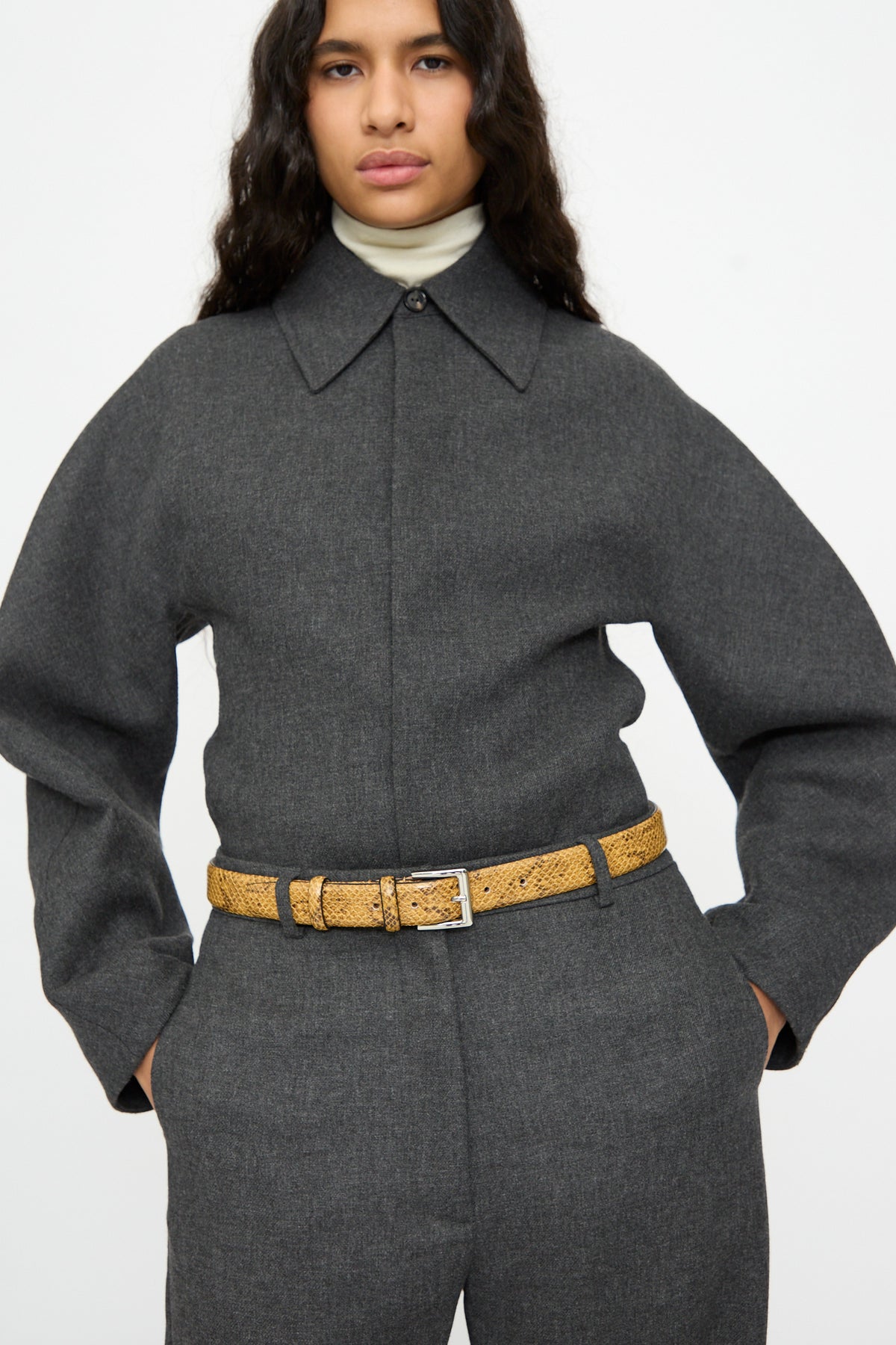 A person wearing a tan Classic Belt in Snake over a charcoal gray collared jumpsuit.
