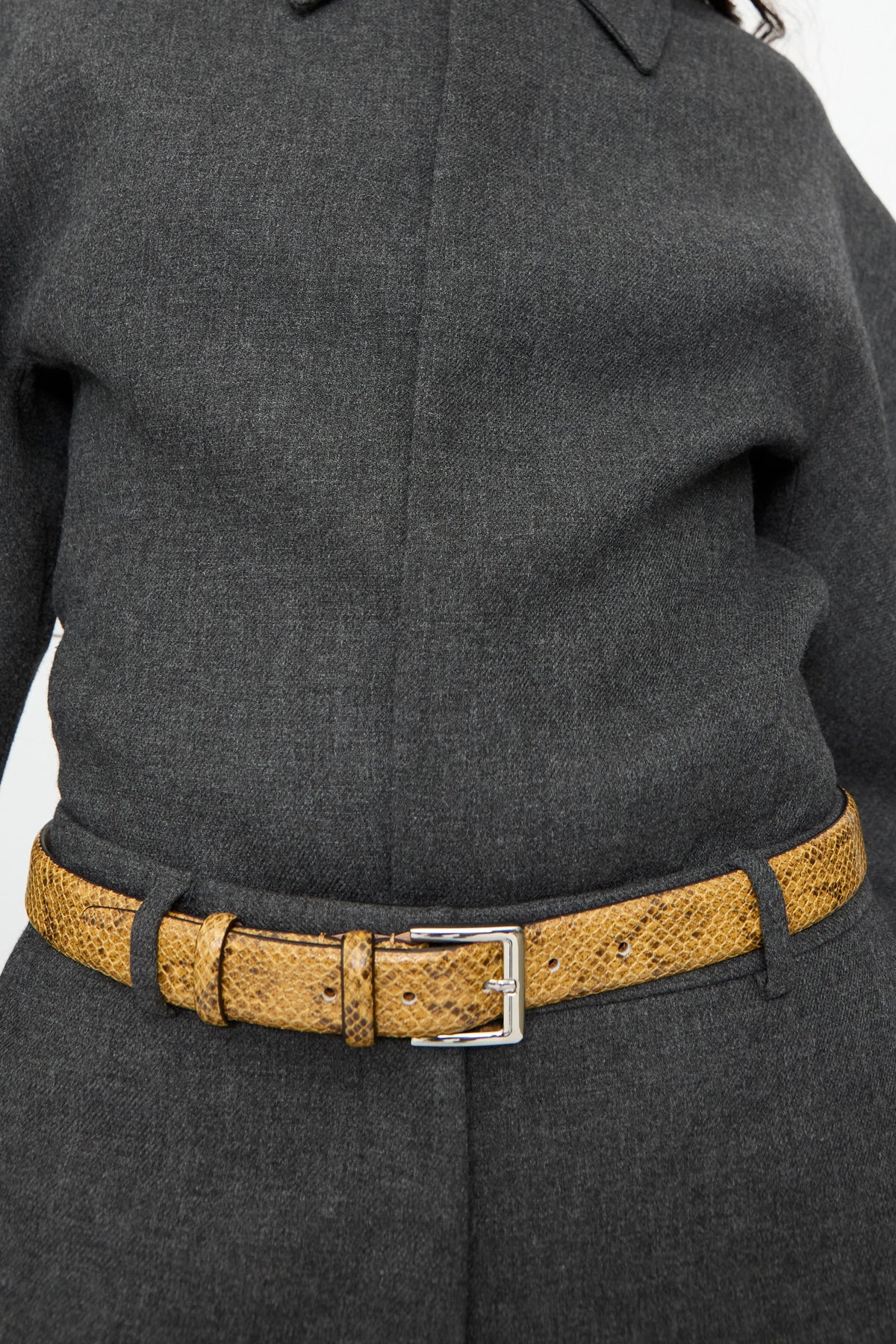 A mustard-yellow Classic Belt in Snake with a silver buckle, worn over a dark grey wool garment.