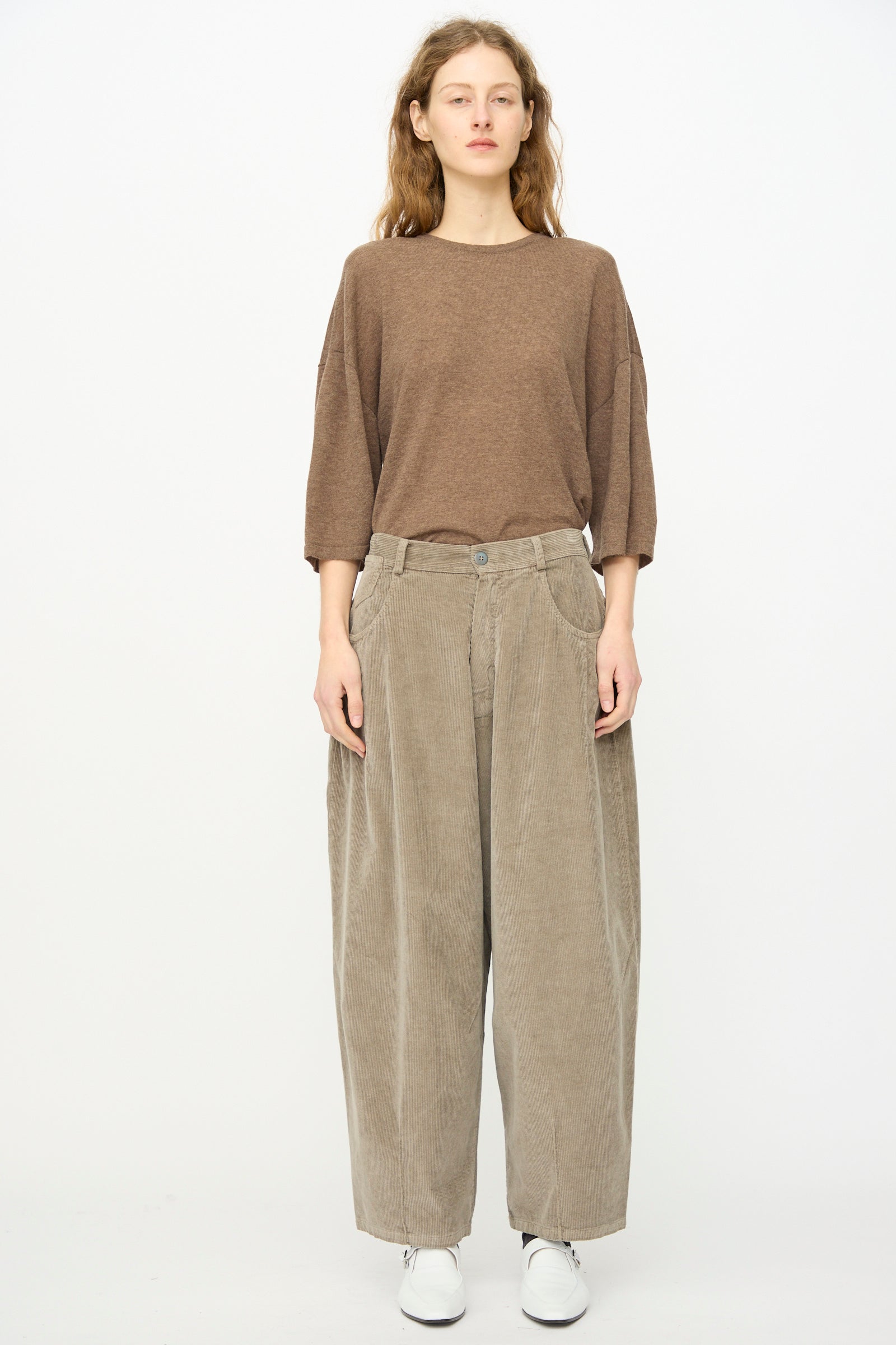 A person stands against a plain background wearing the Cordera Corduroy Baggy Pant in Cobblestone, styled with a loose brown top and white shoes.