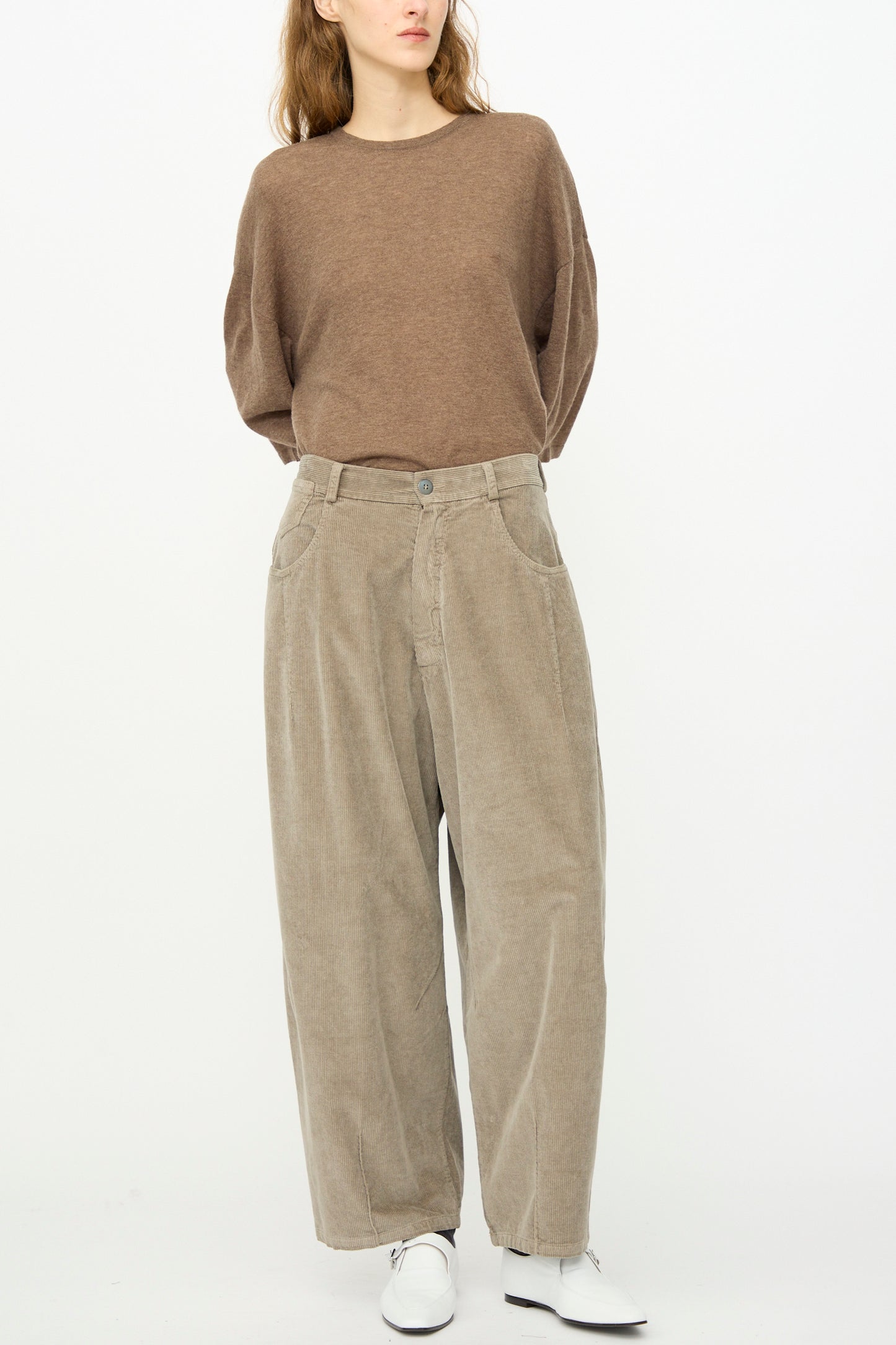 A person wearing Cordera’s Corduroy Baggy Pant in Cobblestone with an elastic waistband, a loose brown long-sleeve top, and white shoes stands against a plain white background.