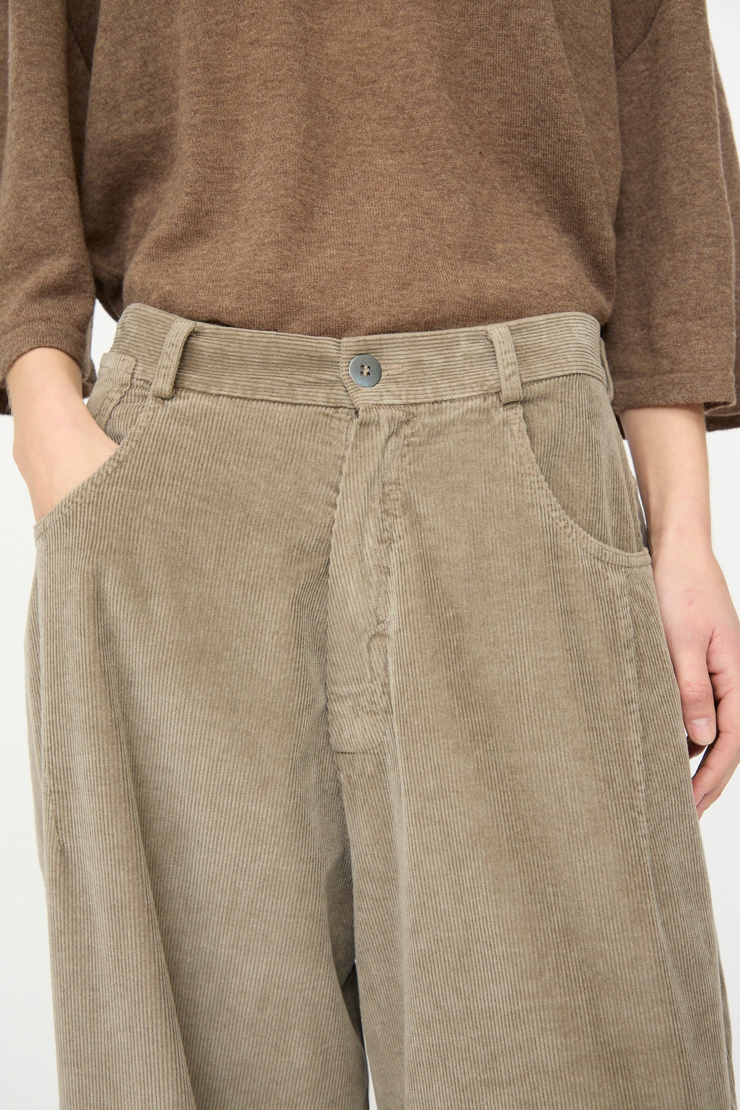 A person wears Cordera’s Corduroy Baggy Pant in Cobblestone, made in Spain with a button closure, paired with a brown top and one hand casually in their pocket.