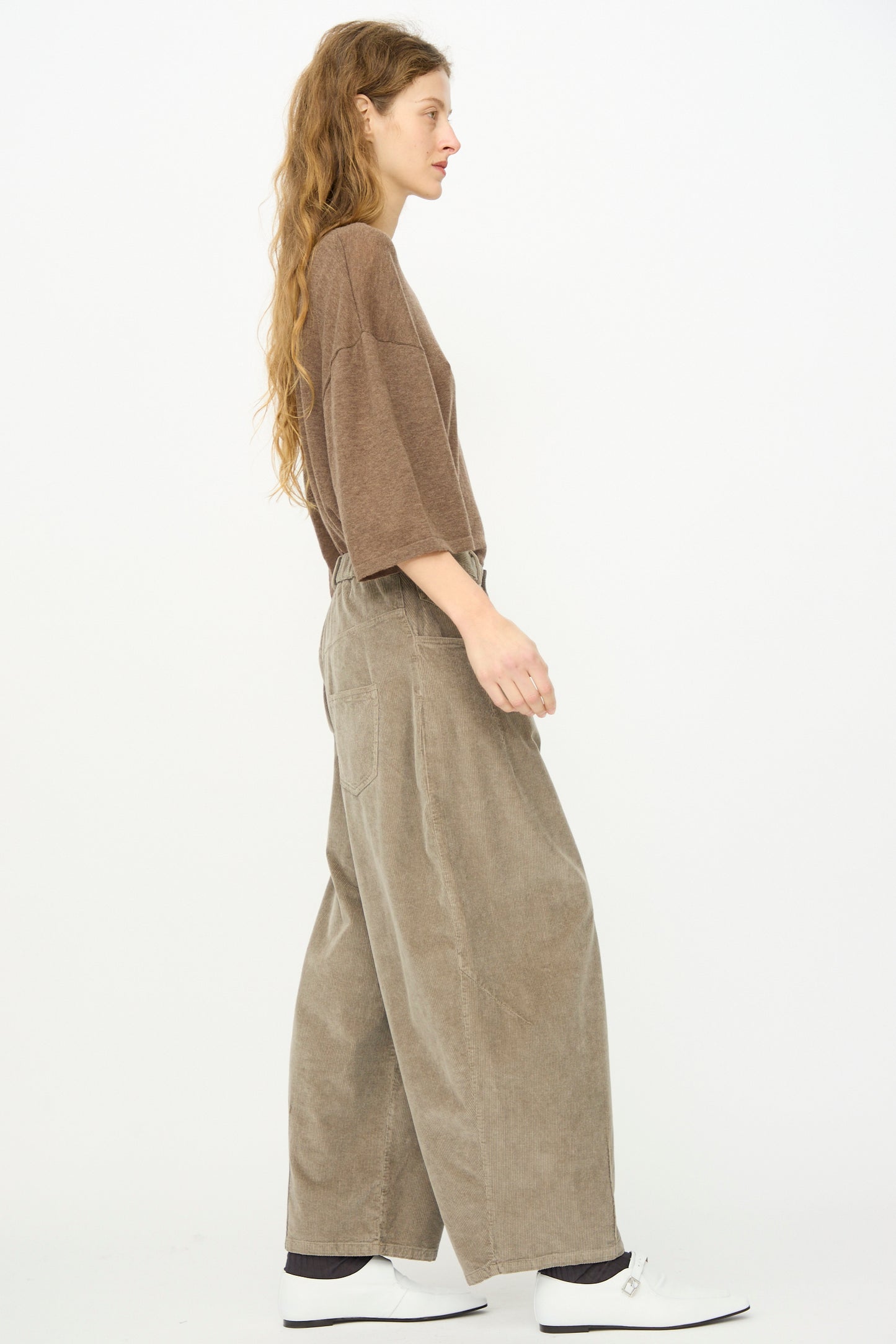 A woman with long hair, wearing a loose brown top, Cordera Corduroy Baggy Pant in Cobblestone with an elastic waistband, and white shoes stands in profile against a plain white background.