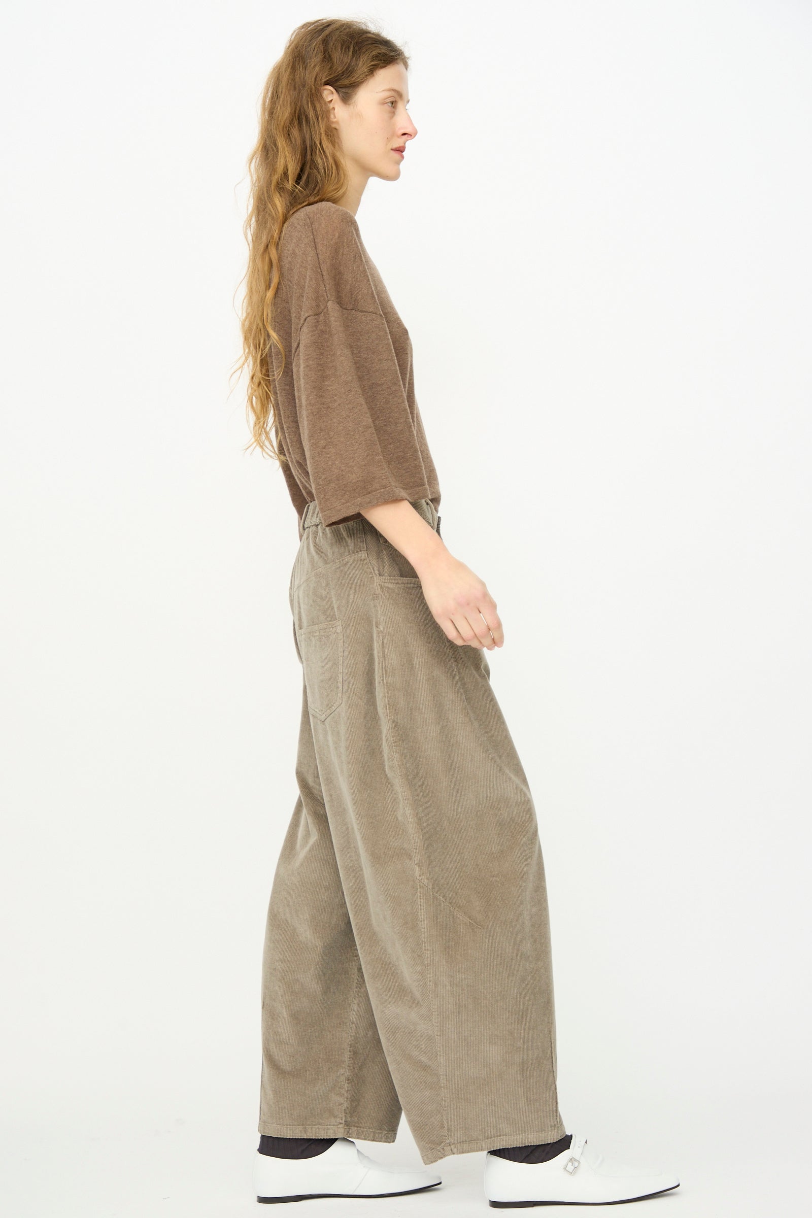 A woman with long hair, wearing a loose brown top, Cordera Corduroy Baggy Pant in Cobblestone with an elastic waistband, and white shoes stands in profile against a plain white background.