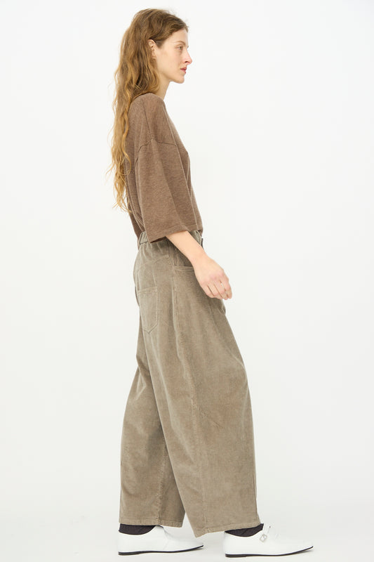 A woman with long hair, wearing a loose brown top, Cordera Corduroy Baggy Pant in Cobblestone with an elastic waistband, and white shoes stands in profile against a plain white background.