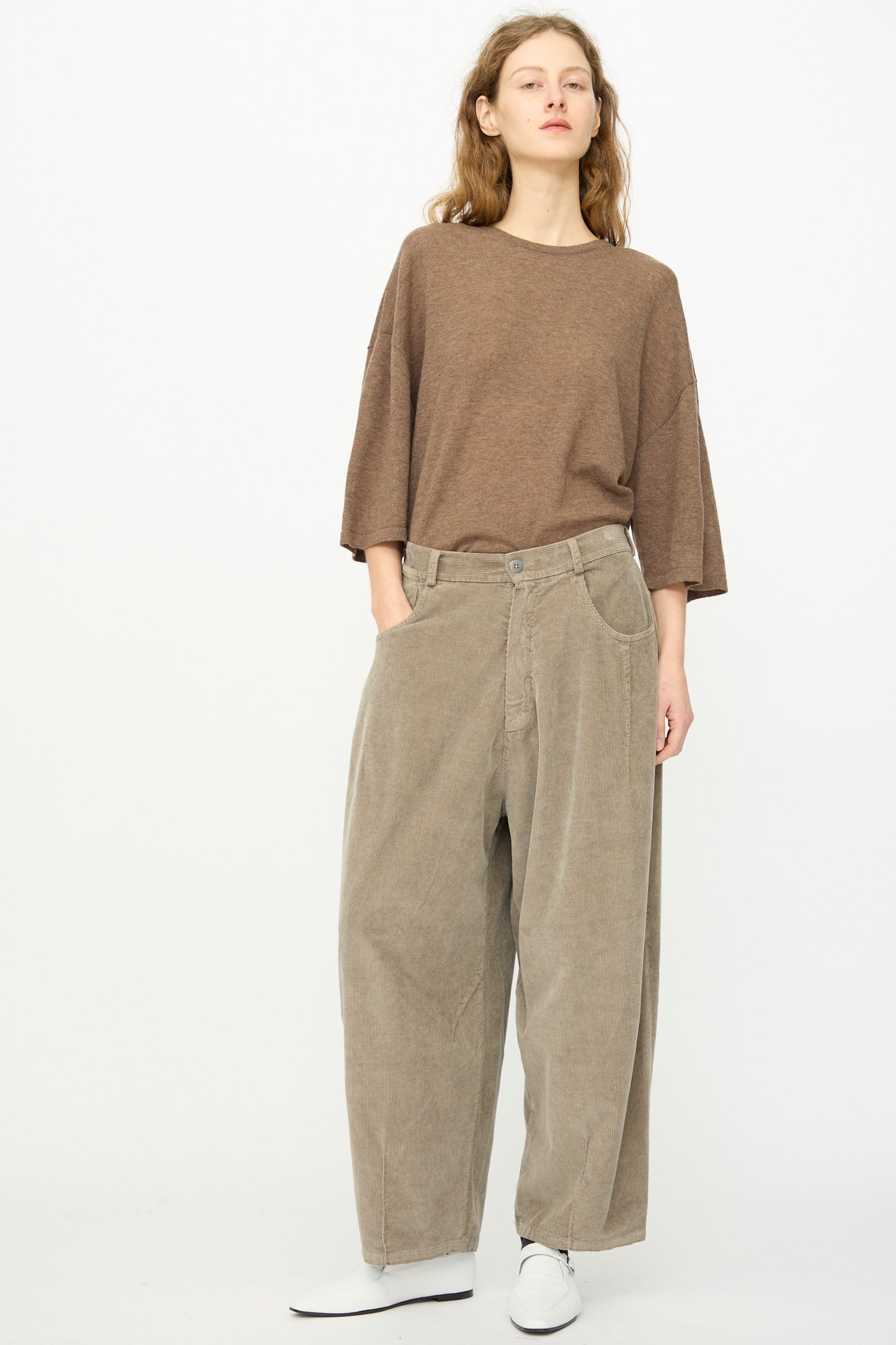 A person stands against a plain background wearing Cordera's Corduroy Baggy Pant in Cobblestone, styled with a loose brown top and white shoes, hands in pockets.