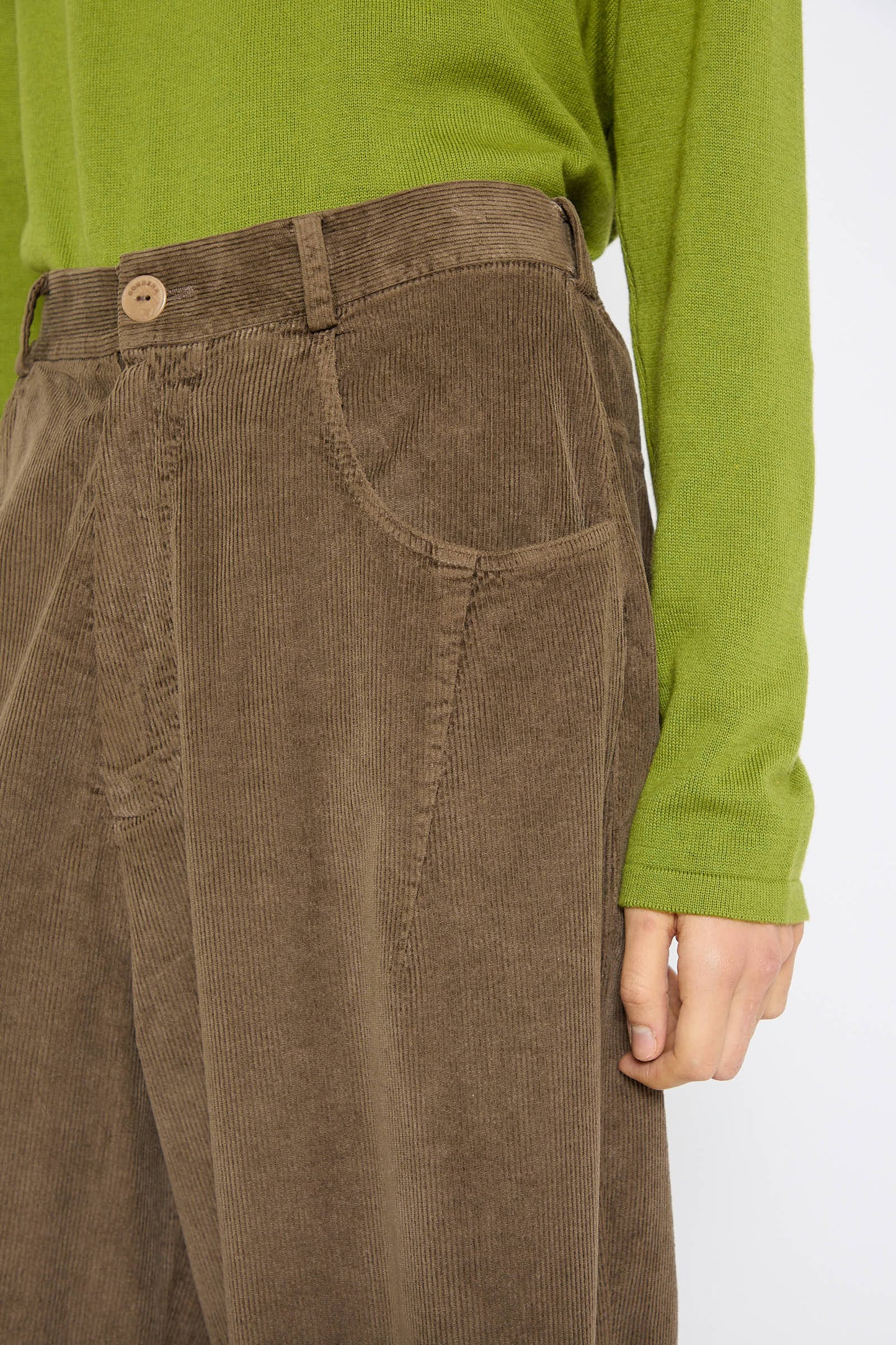 Close-up of a person wearing Cordera's Corduroy Baggy Pant in Moss with a green long-sleeve shirt. The focus is on the waistband and pocket area of the pants.