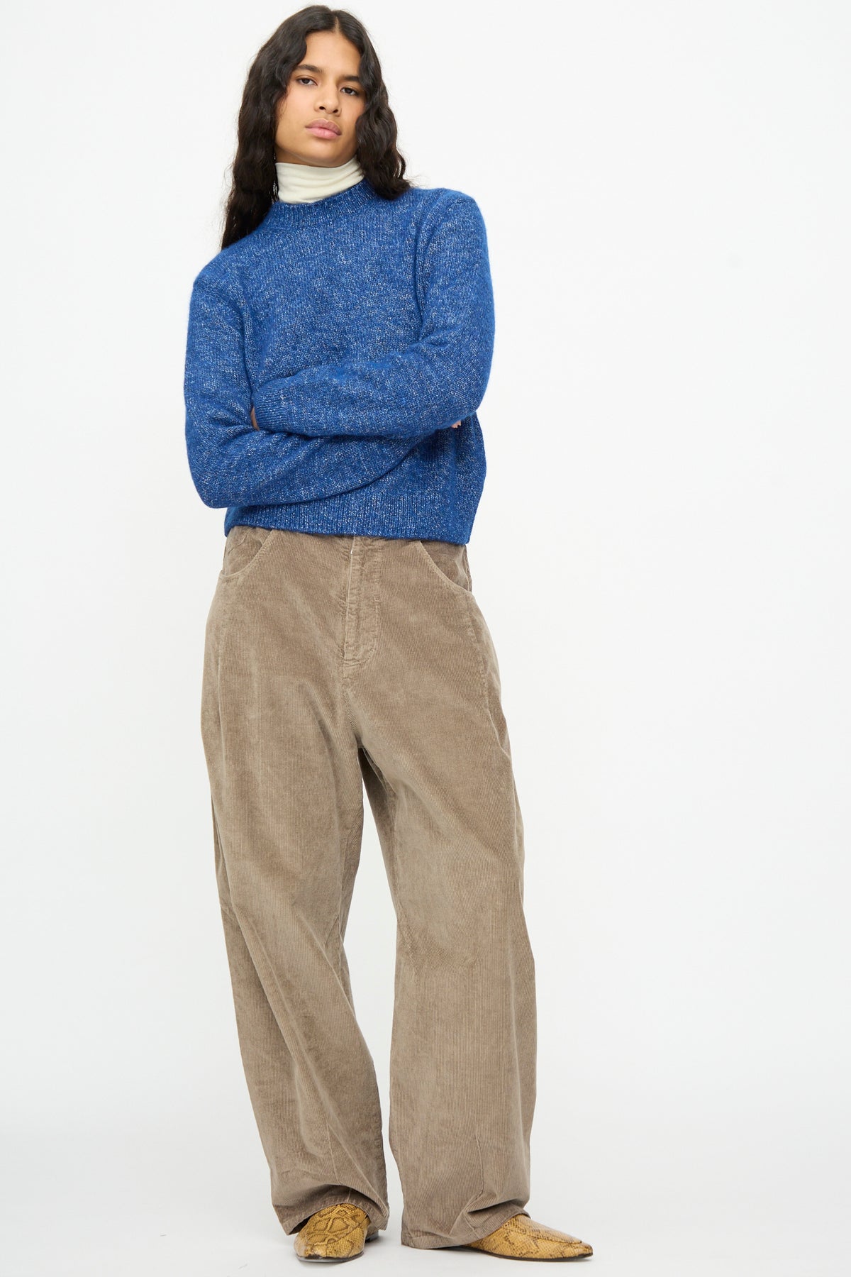A person with arms crossed wears a blue knit sweater over a cream turtleneck and Cordera Corduroy Baggy Pants in Sage.