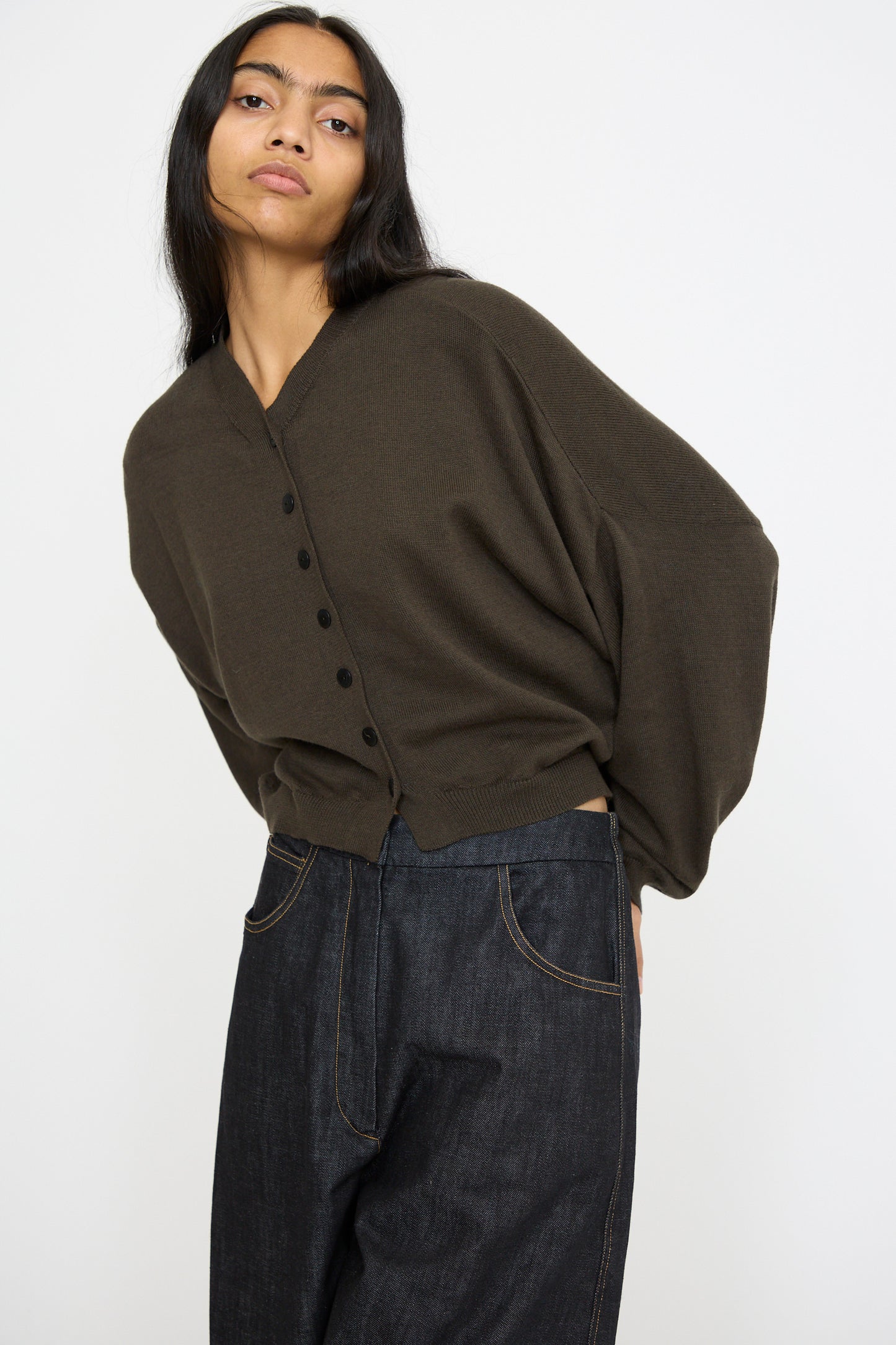 A person stands against a plain background wearing Cordera's Cotton and Cashmere Cardigan in Dark Moss, paired with dark wide-leg pants. 