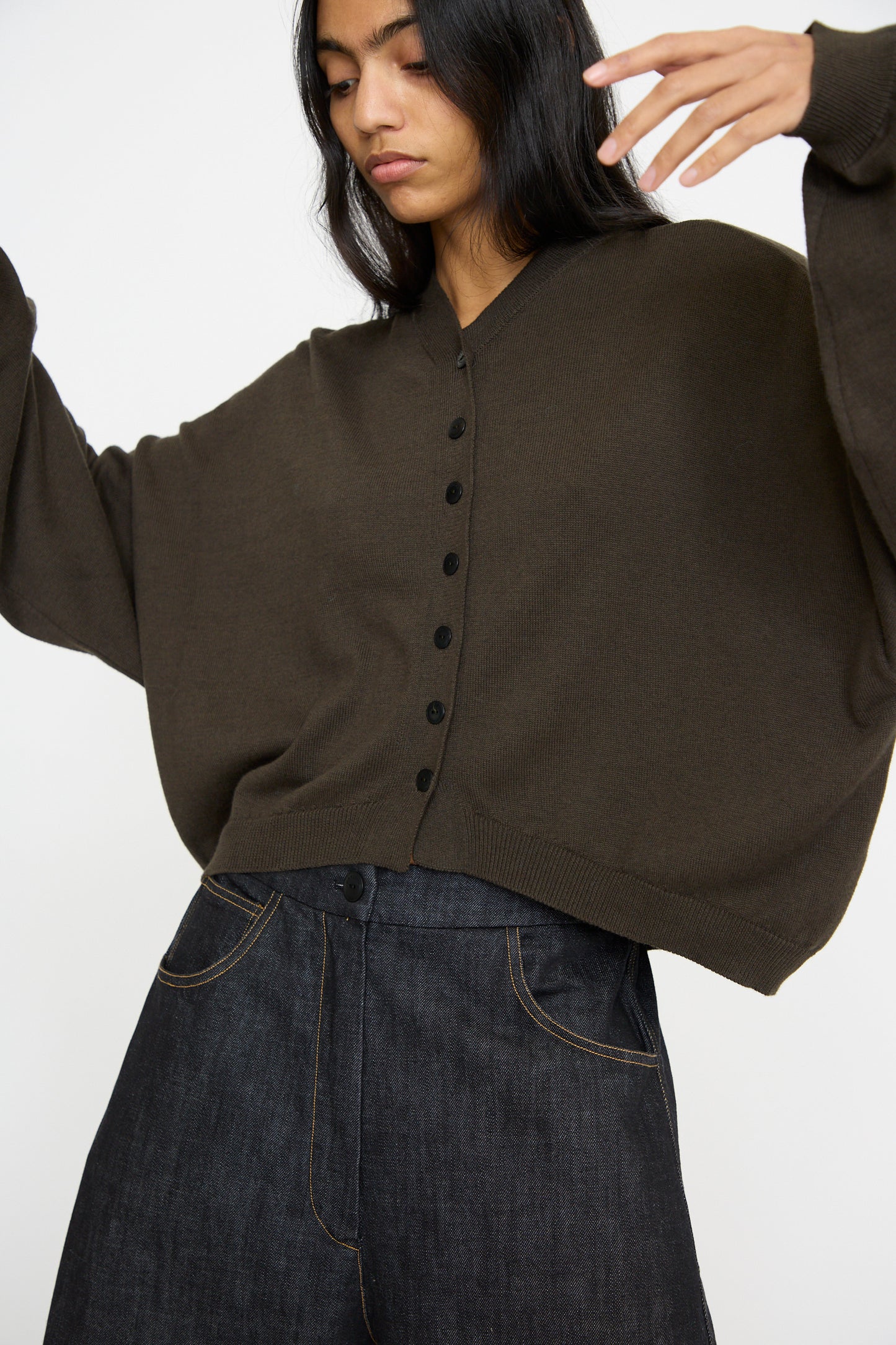 Someone wearing a loose fit Cordera Cotton and Cashmere Cardigan in Dark Moss with black buttons and dark denim pants, posing against a plain background.