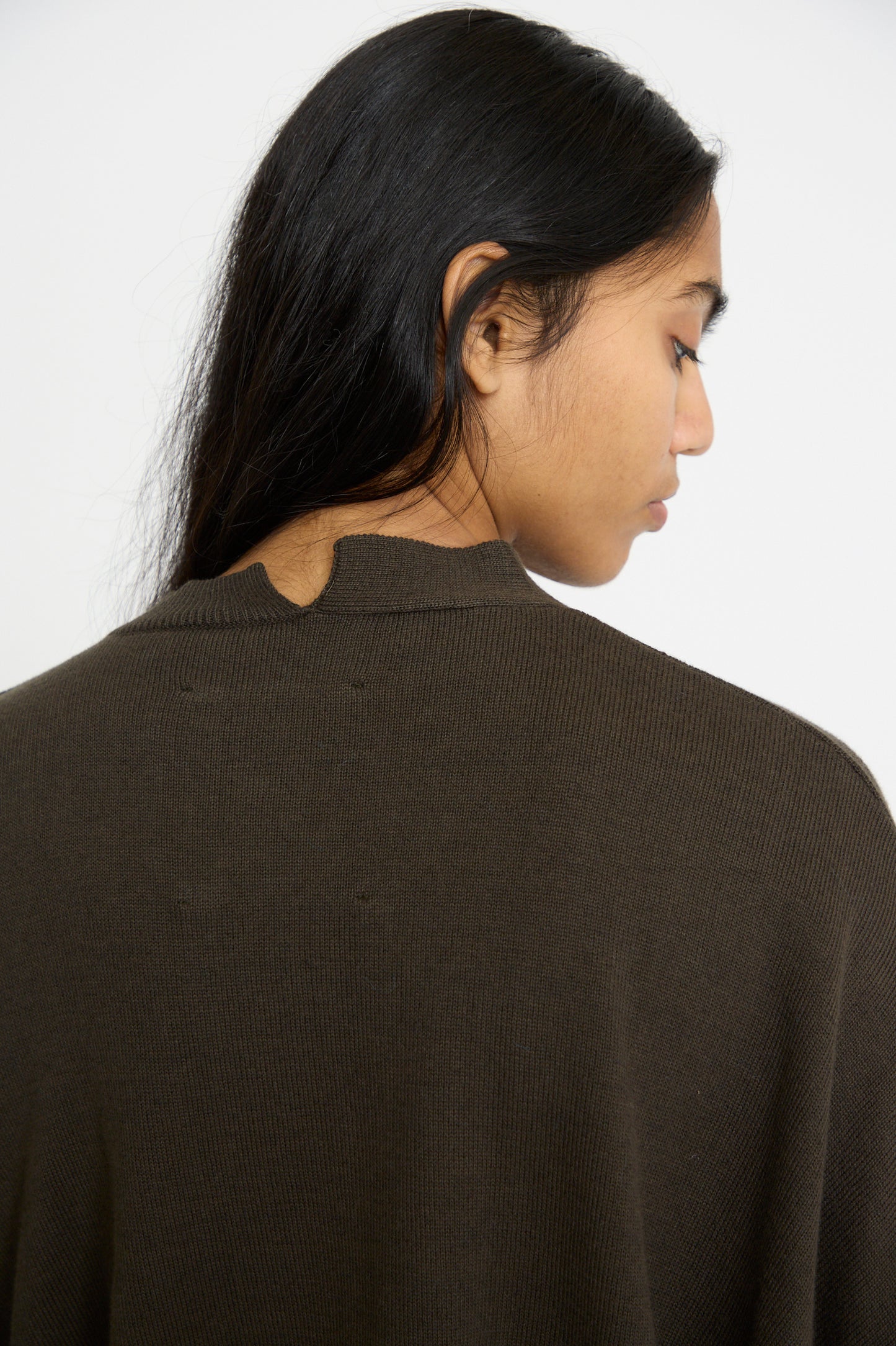 A person with long dark hair is shown from the side and back, wearing Cordera's Cotton and Cashmere Cardigan in Dark Moss, against a white background. 