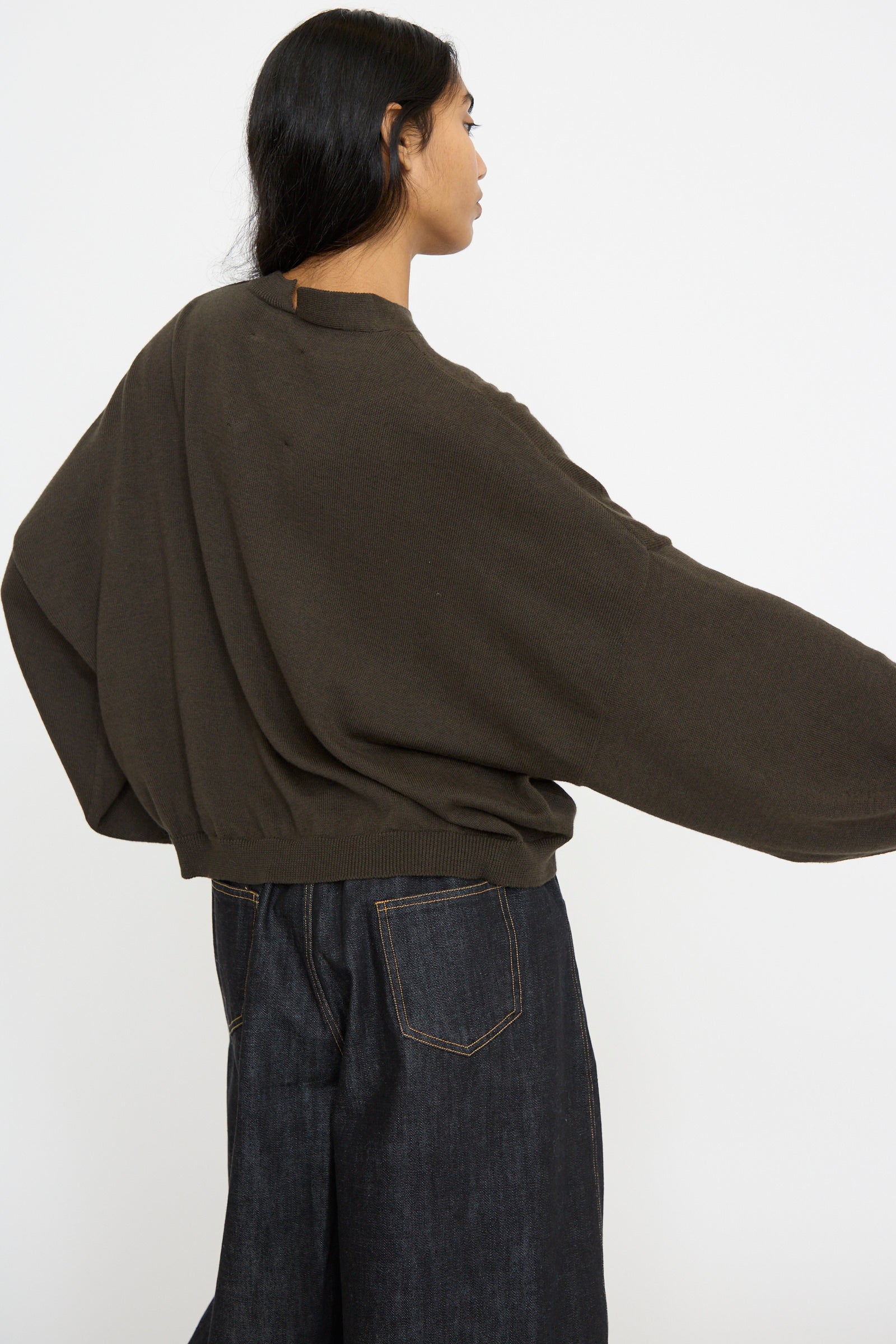 A person wearing an oversized dark moss cotton and cashmere blend cardigan by Cordera with dark jeans poses relaxedly to the left against a plain background.