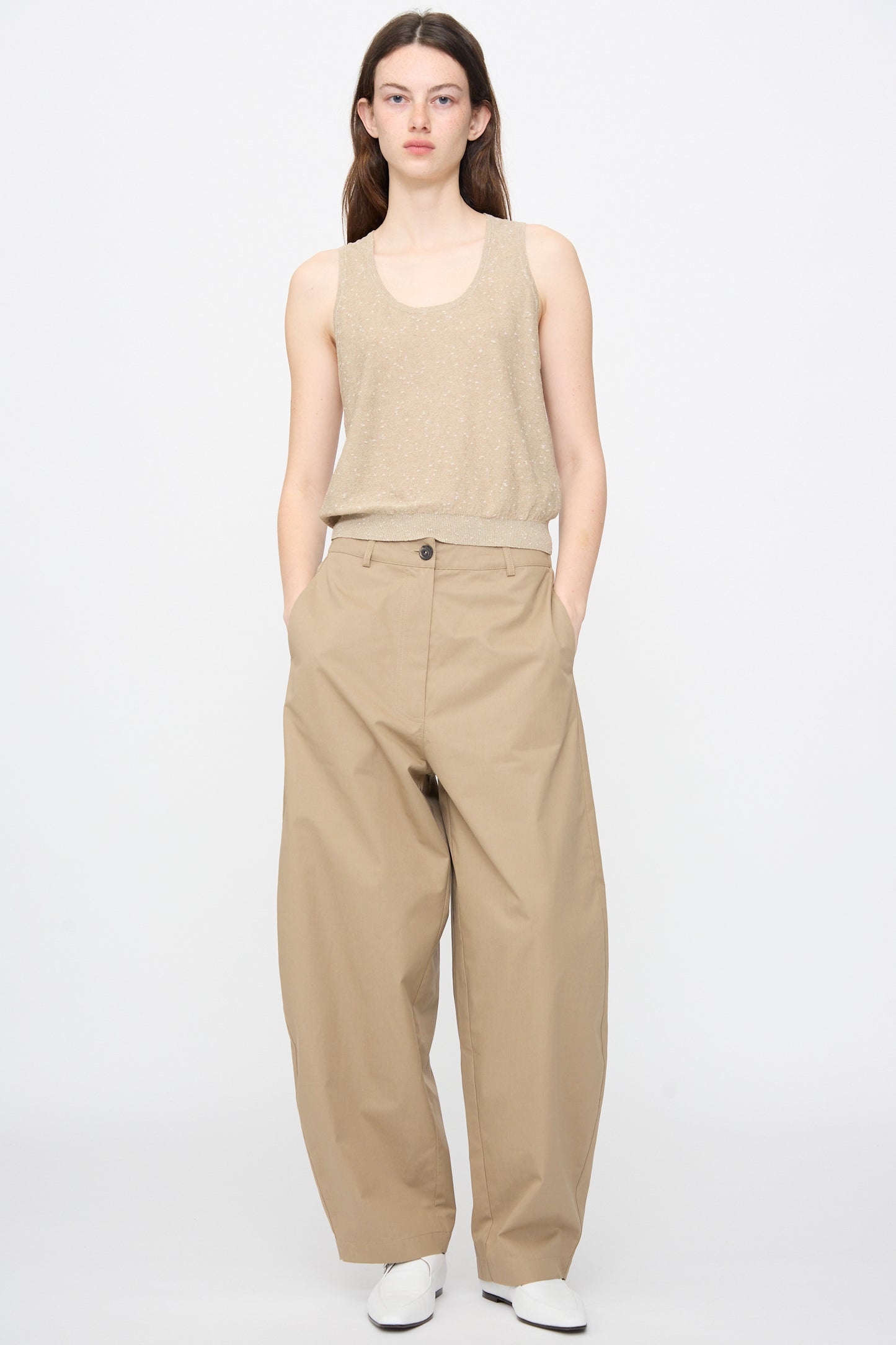 A person stands against a plain background wearing Cordera's Cotton Curved Pants in Camel, paired with a beige sleeveless top and white shoes, hands in pockets.