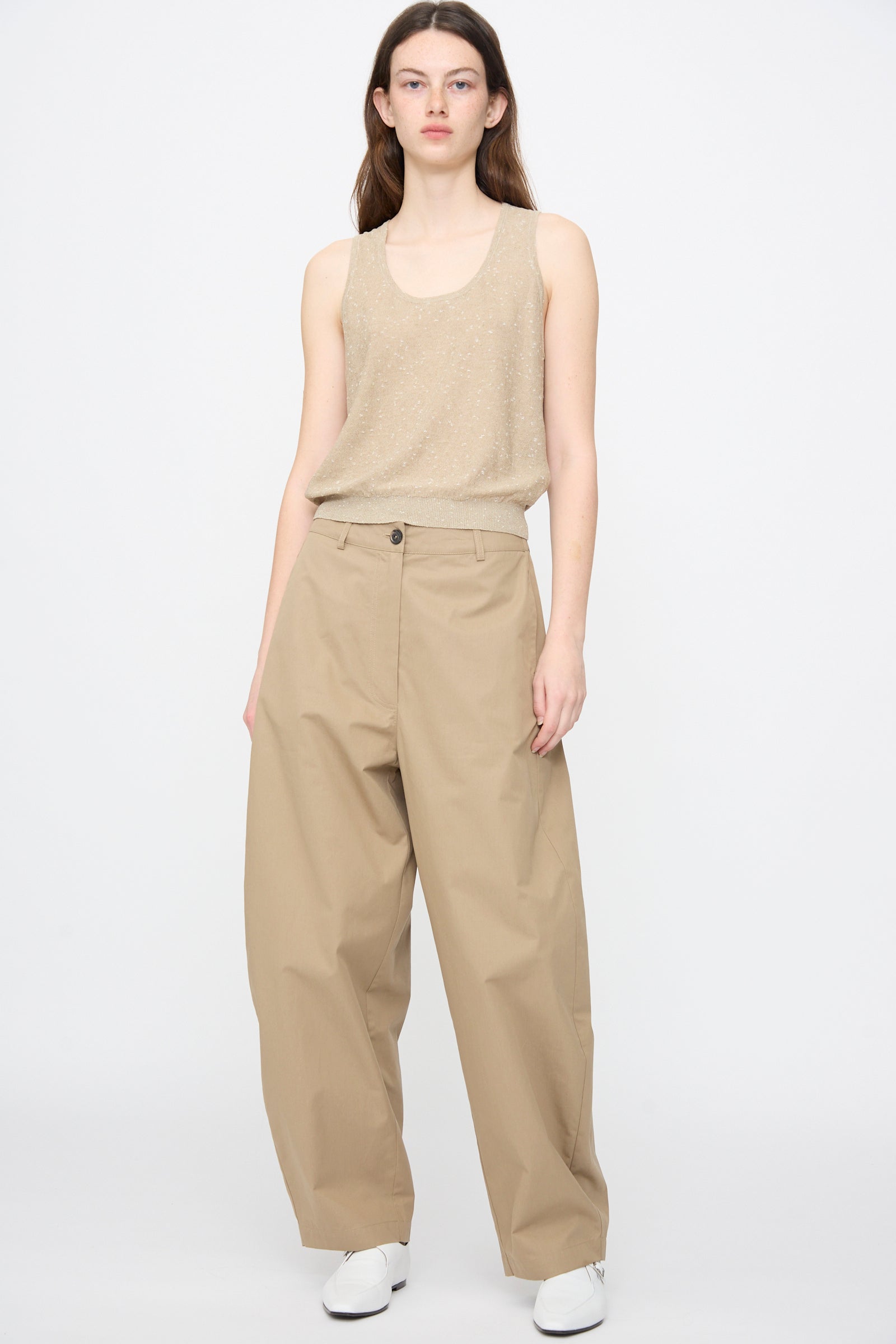 A person wearing a beige sleeveless top, Cordera Cotton Curved Pants in Camel, and white shoes stands against a plain white background.