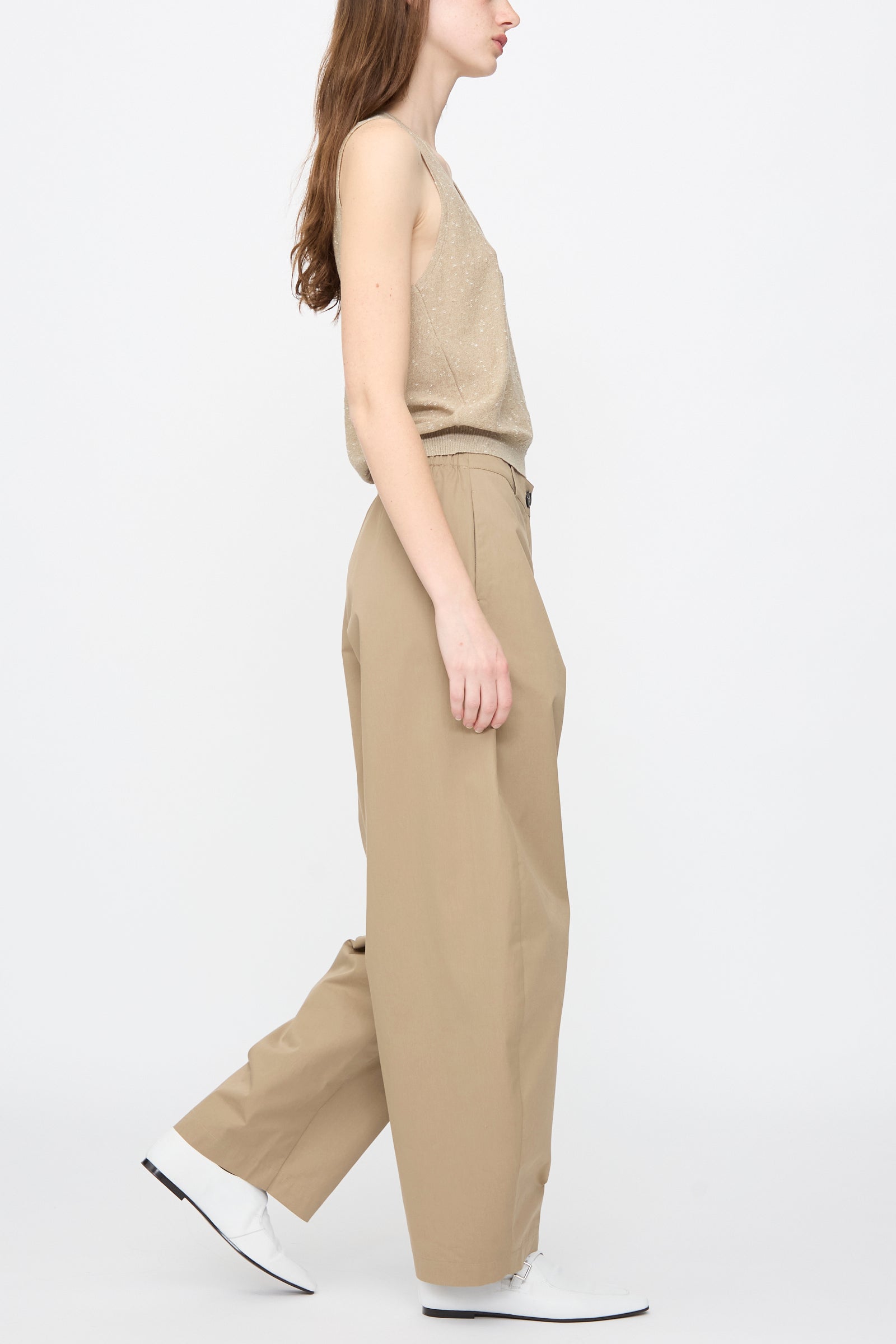 A person wearing Cordera’s Cotton Curved Pants in Camel and a sleeveless beige top walks in profile against a plain white background.