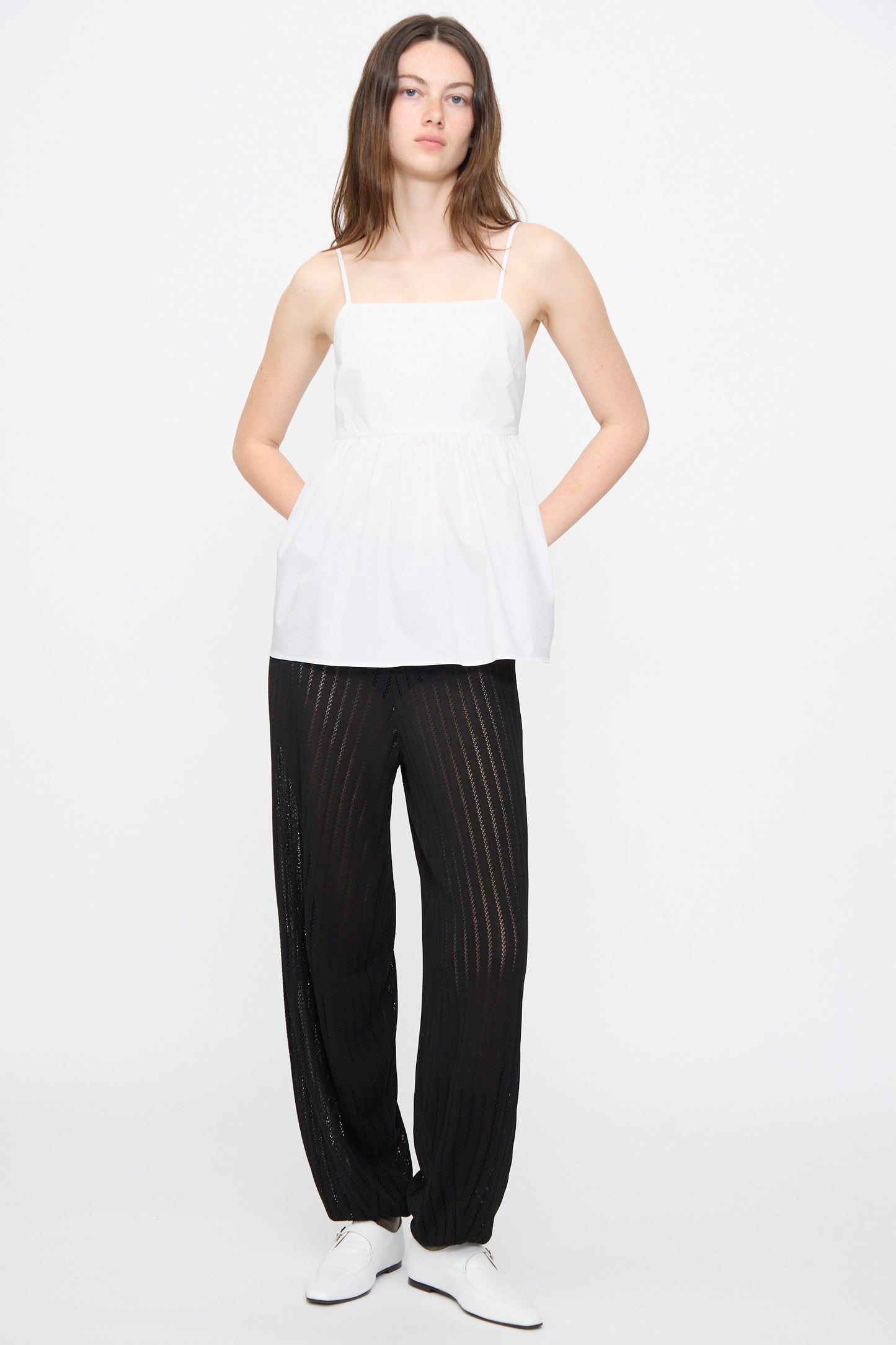 A woman stands against a white background wearing the Cordera Cotton Embroidered Top in White, paired with black sheer pants and white shoes, her hands tucked in her pockets.