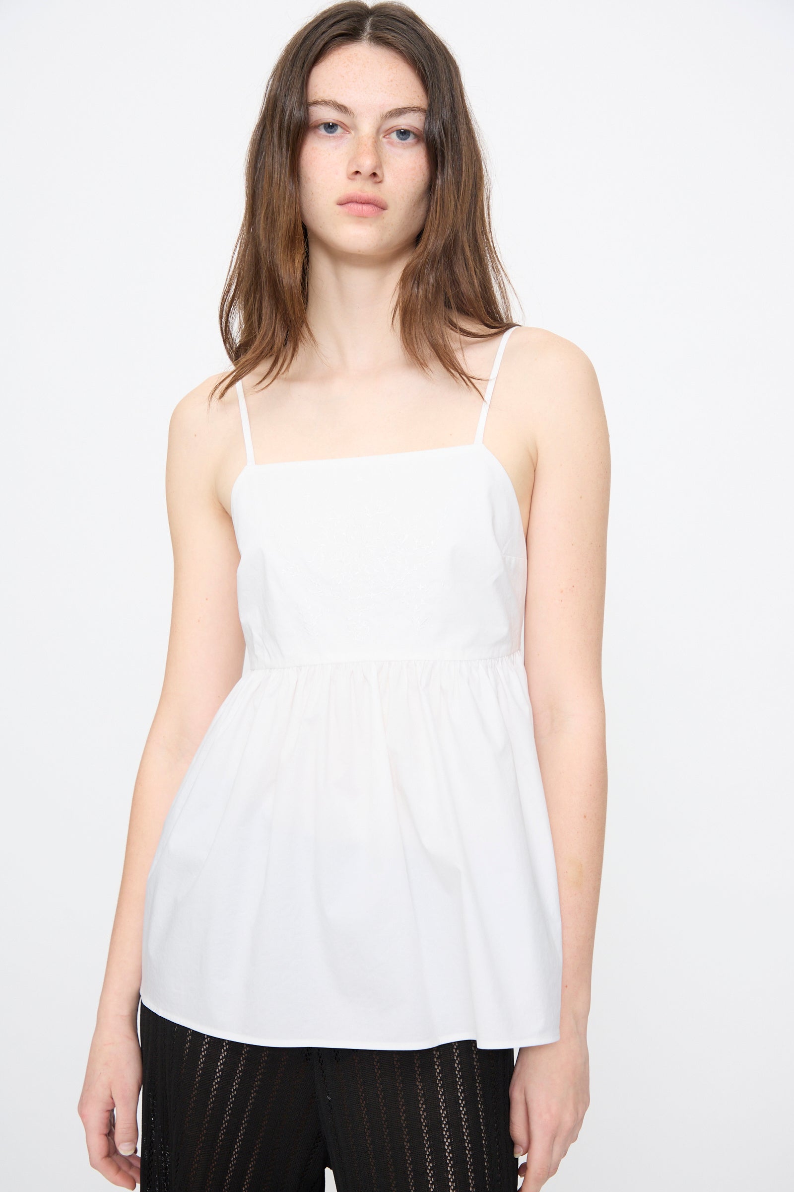 A woman with long brown hair models the Cordera Cotton Embroidered Top in White, featuring an adjustable back tie, paired with black pants against a plain white background.