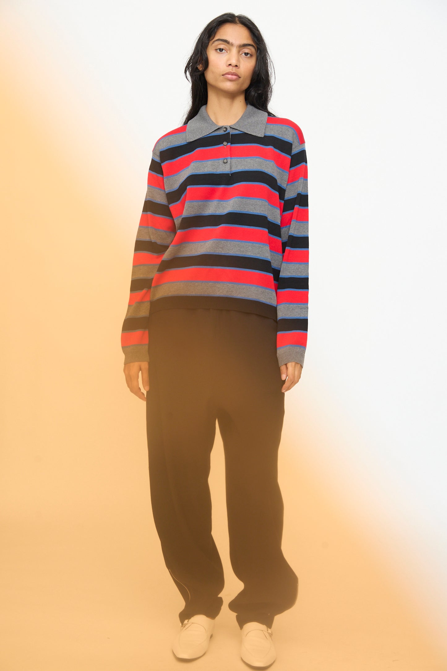 A person stands against a plain background, wearing the Cordera Cotton Long Sleeve Polo in Stripe featuring an oversized fit with red, navy, and gray stripes, paired with loose black pants and white shoes. A yellowish light effect appears on the left side.
