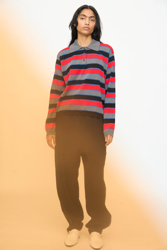 A person stands against a plain background, wearing the Cordera Cotton Long Sleeve Polo in Stripe featuring an oversized fit with red, navy, and gray stripes, paired with loose black pants and white shoes. A yellowish light effect appears on the left side.