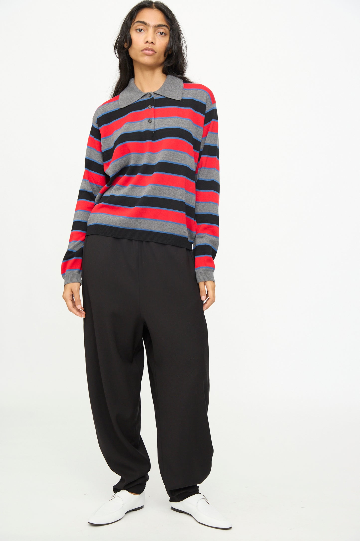 A person stands against a plain background wearing Cordera’s Cotton Long Sleeve Polo in Stripe—an oversized-fit gray and red striped polo—paired with loose black pants and white shoes.