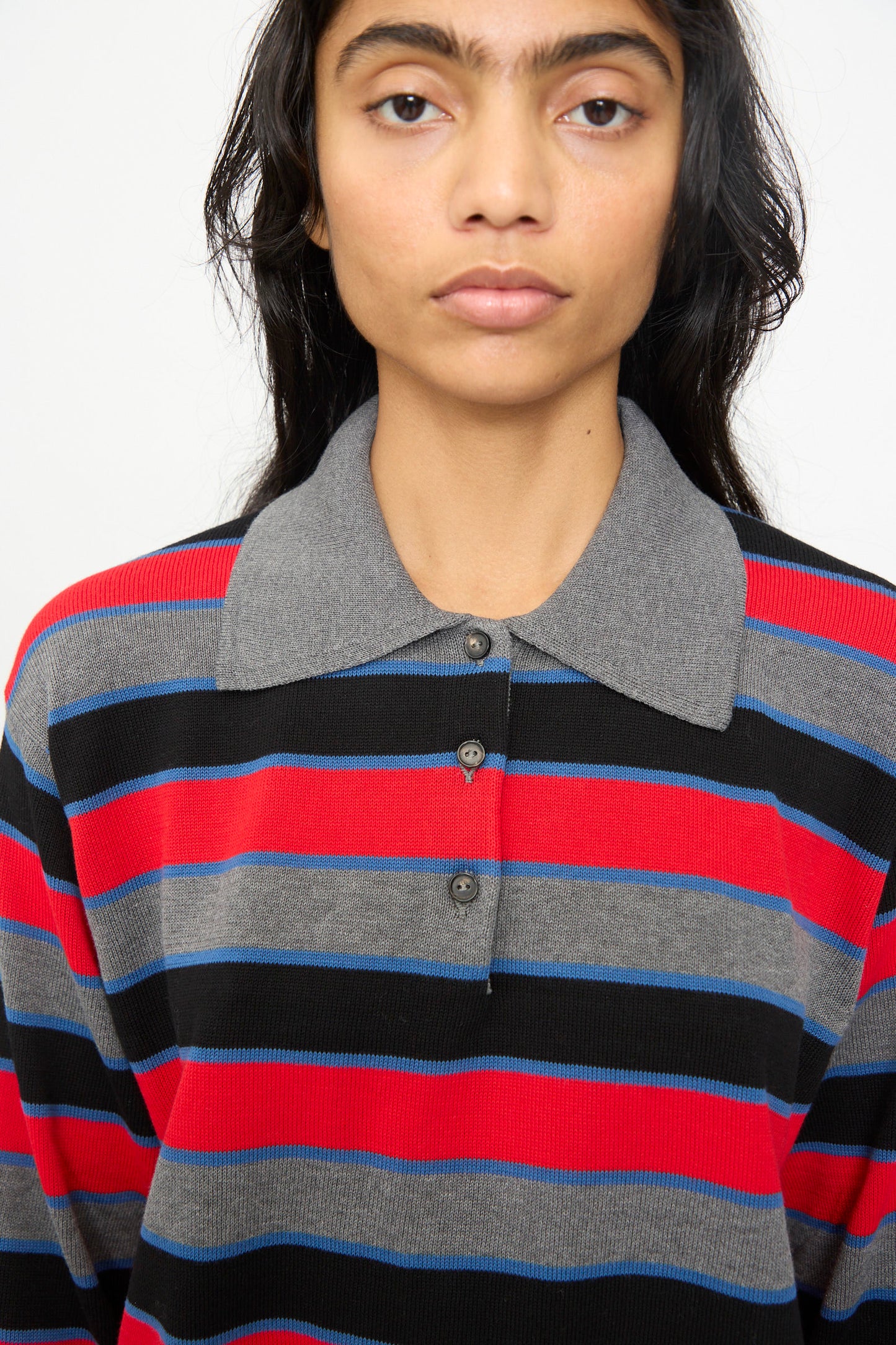 A person with long dark hair faces the camera against a plain background, wearing Cordera's Cotton Long Sleeve Polo in Stripe, an oversized fit with a gray collar and red, black, and gray horizontal stripes.