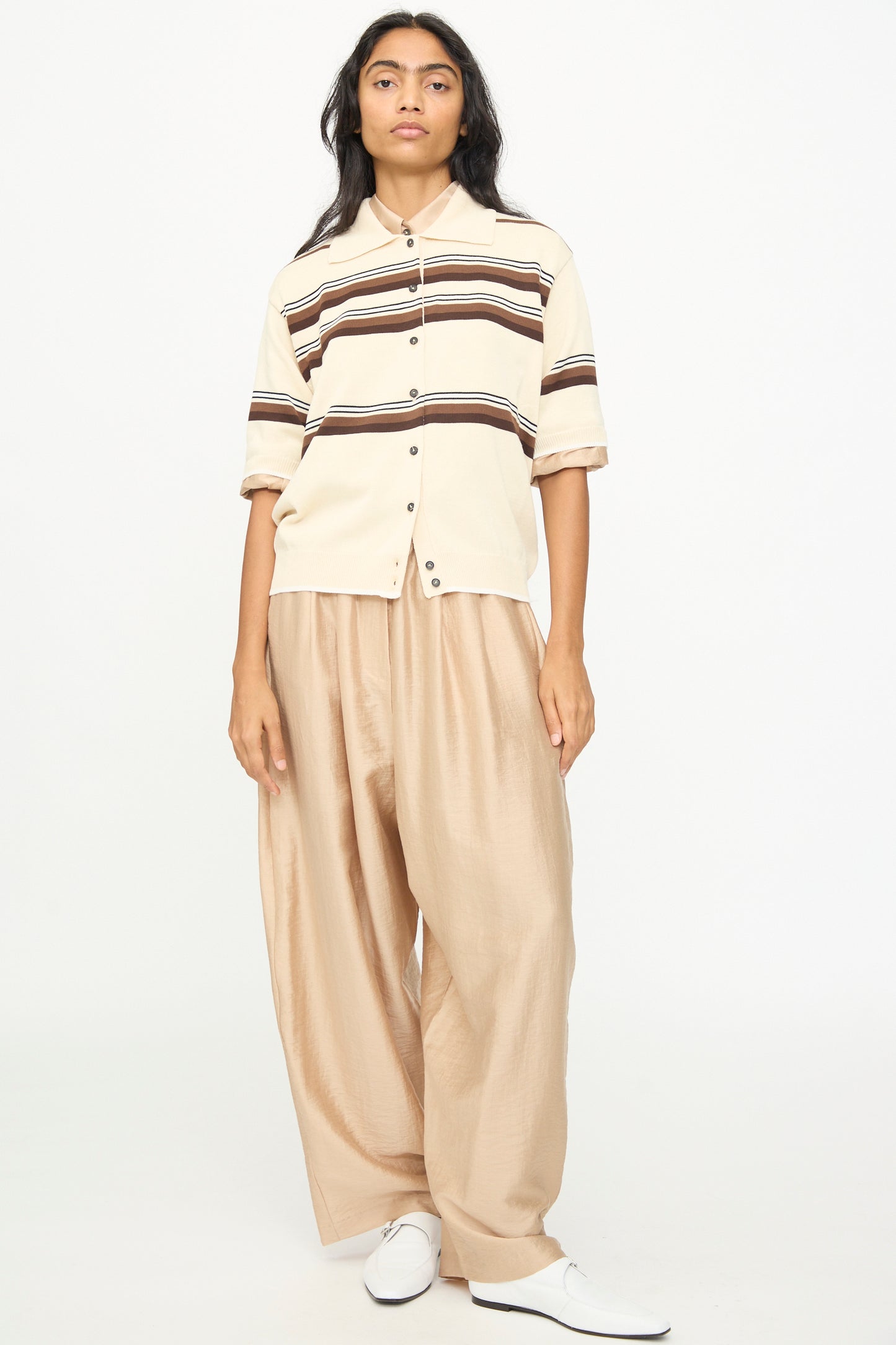 A person is seen standing against a plain white background, wearing the Cordera Cotton Polo in Striped—a beige short sleeve shirt with brown stripes and a polo collar—paired with loose tan pants and white shoes.