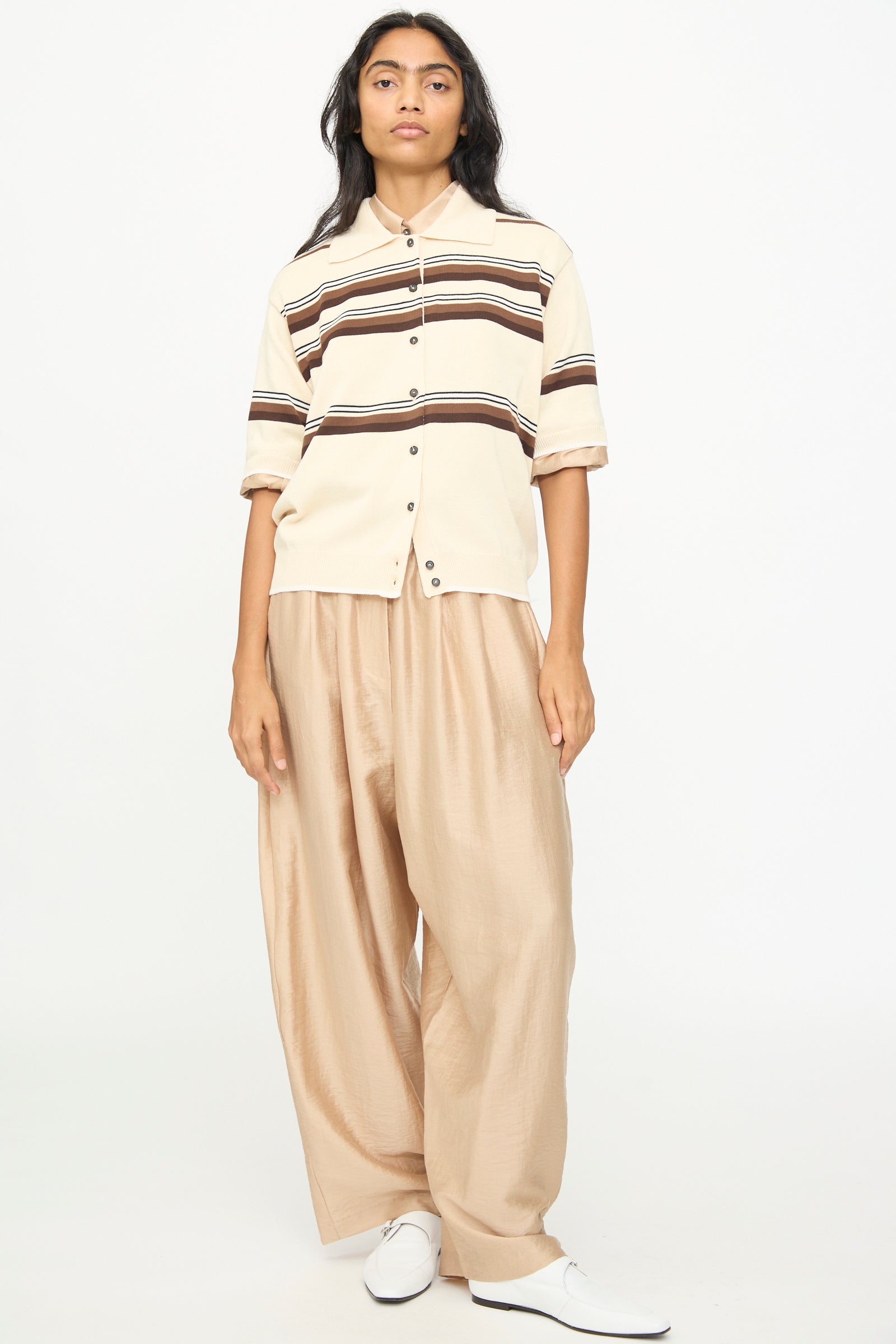 A person is seen standing against a plain white background, wearing the Cordera Cotton Polo in Striped—a beige short sleeve shirt with brown stripes and a polo collar—paired with loose tan pants and white shoes.