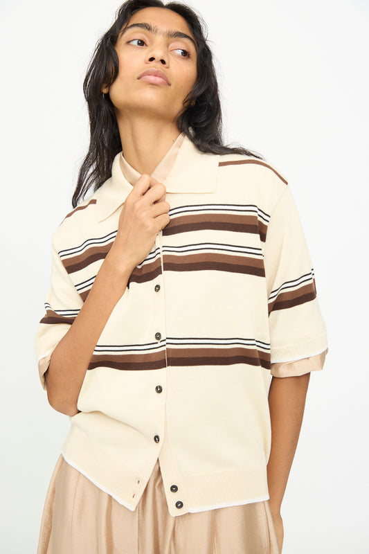 A person models the Cordera Cotton Polo in Striped—a cream, short-sleeve cotton shirt with brown horizontal stripes and a polo collar—paired with a beige skirt, holding the collar and gazing to the side against a plain background.