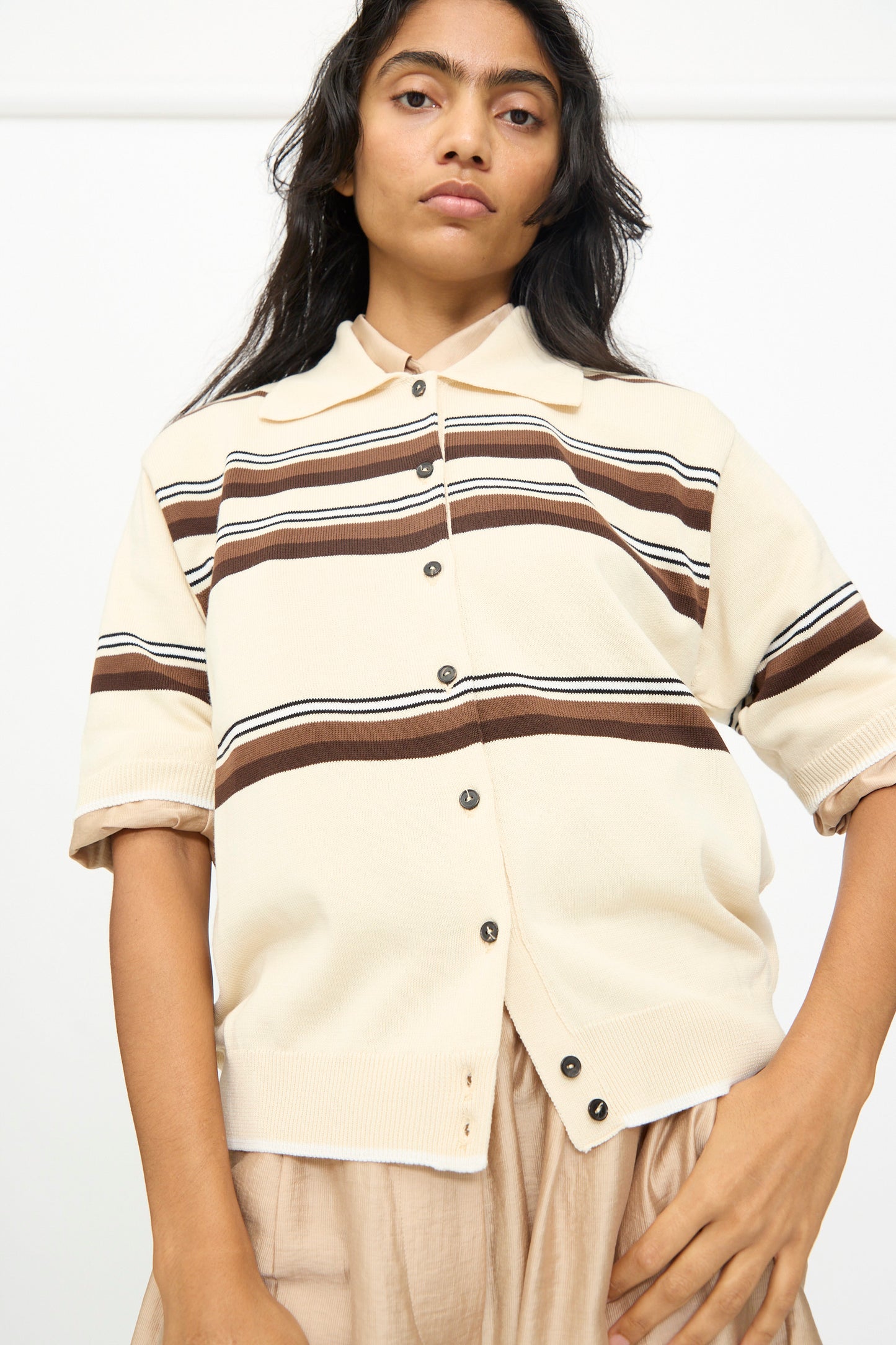 A woman is wearing the Cordera Cotton Polo in Striped—a beige short-sleeved button-up sweater featuring brown horizontal stripes and a classic polo collar—standing against a plain white background.