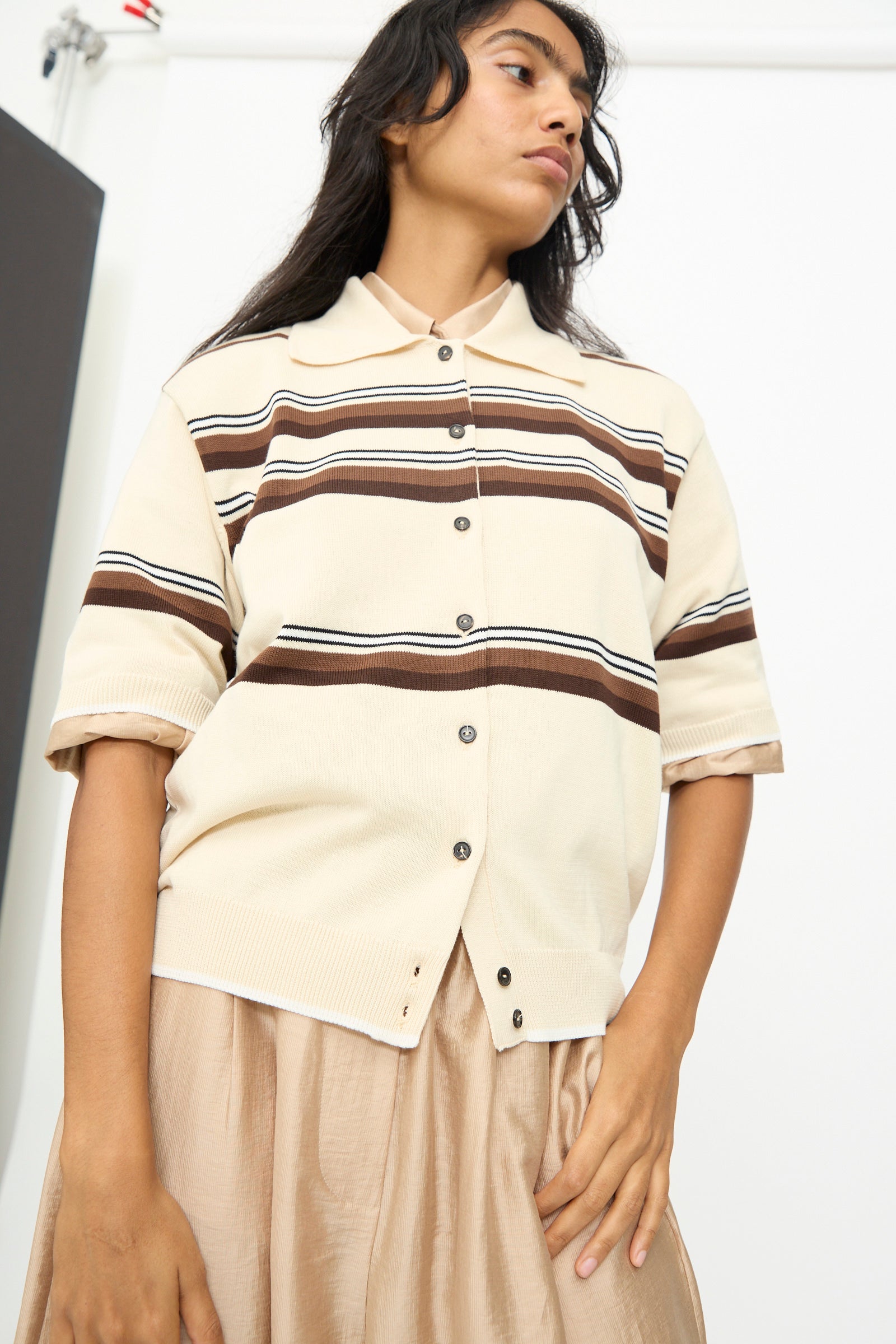 A person is wearing the Cordera Cotton Polo in Striped—a beige short sleeve cotton shirt with a polo collar and brown horizontal stripes—paired with a beige skirt, standing indoors against a plain background.