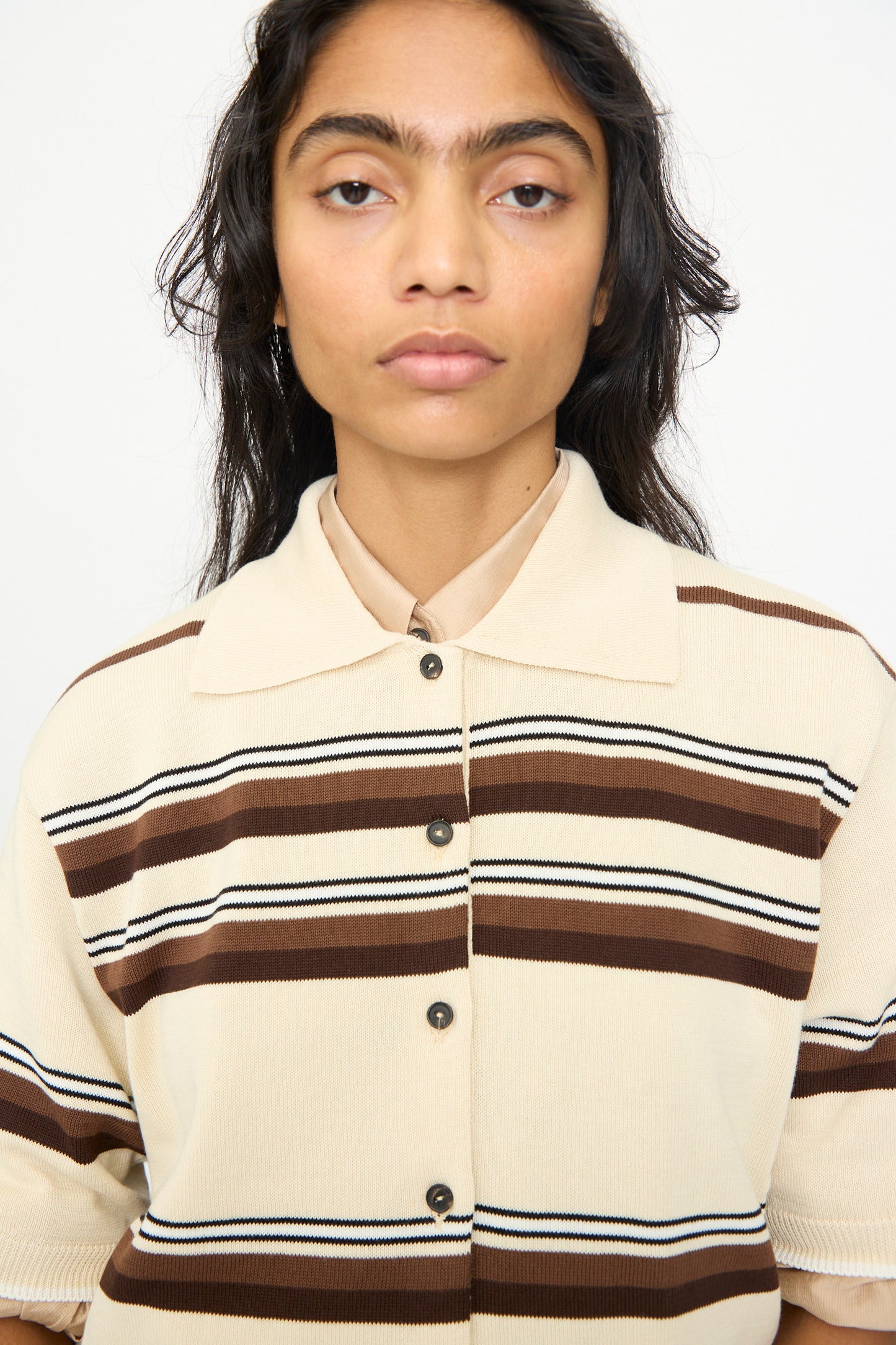 A person with long dark hair is wearing the Cordera Cotton Polo in Striped—a beige shirt featuring brown and black horizontal stripes—while standing against a plain white background.