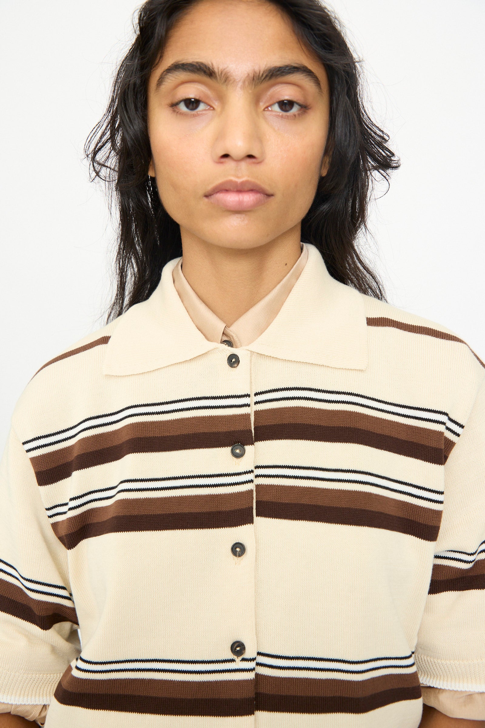 A person with long dark hair is wearing the Cordera Cotton Polo in Striped—a beige shirt featuring brown and black horizontal stripes—while standing against a plain white background.
