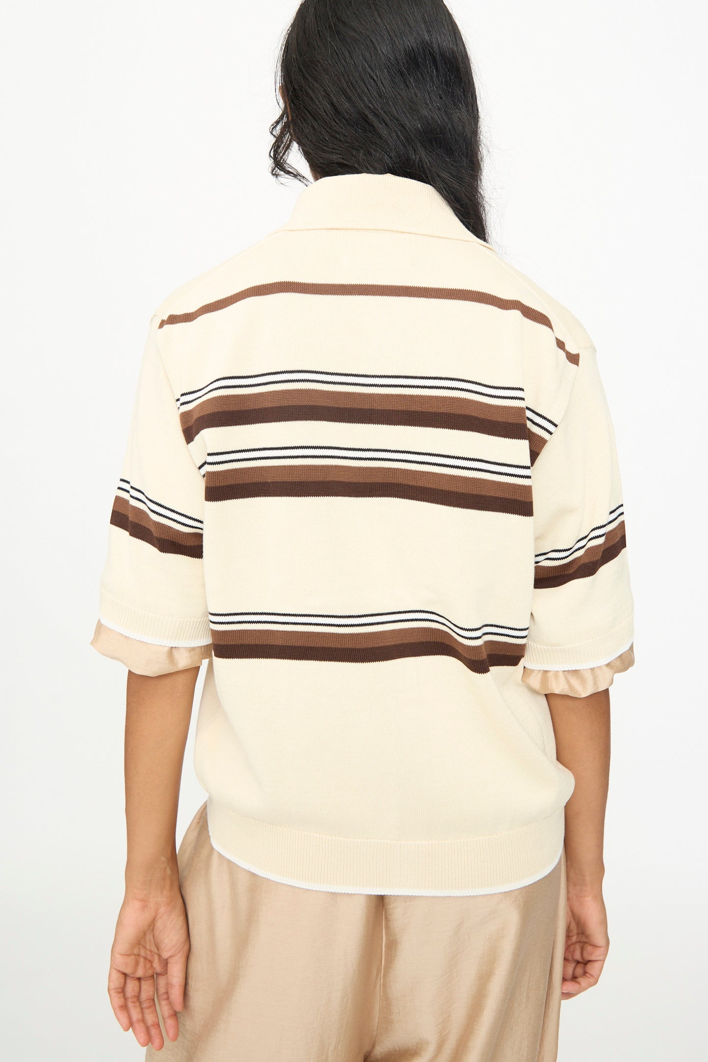 A person stands with their back to the camera, wearing the Cordera Cotton Polo in Striped—a cream-colored short sleeve cotton shirt featuring brown horizontal stripes—paired with light brown pants.