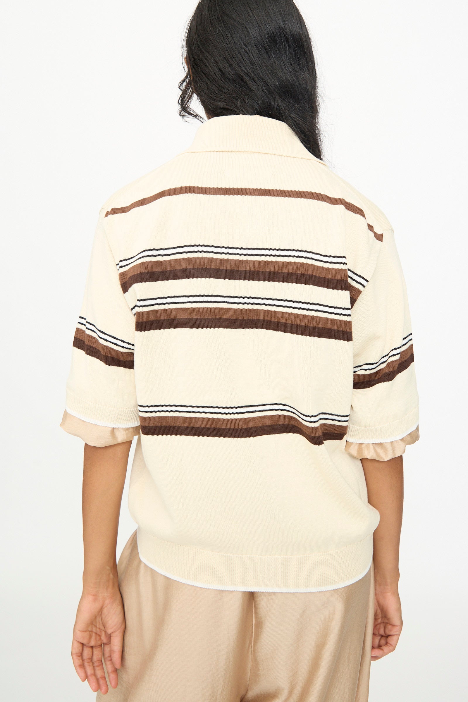 A person stands with their back to the camera, wearing the Cordera Cotton Polo in Striped—a cream-colored short sleeve cotton shirt featuring brown horizontal stripes—paired with light brown pants.